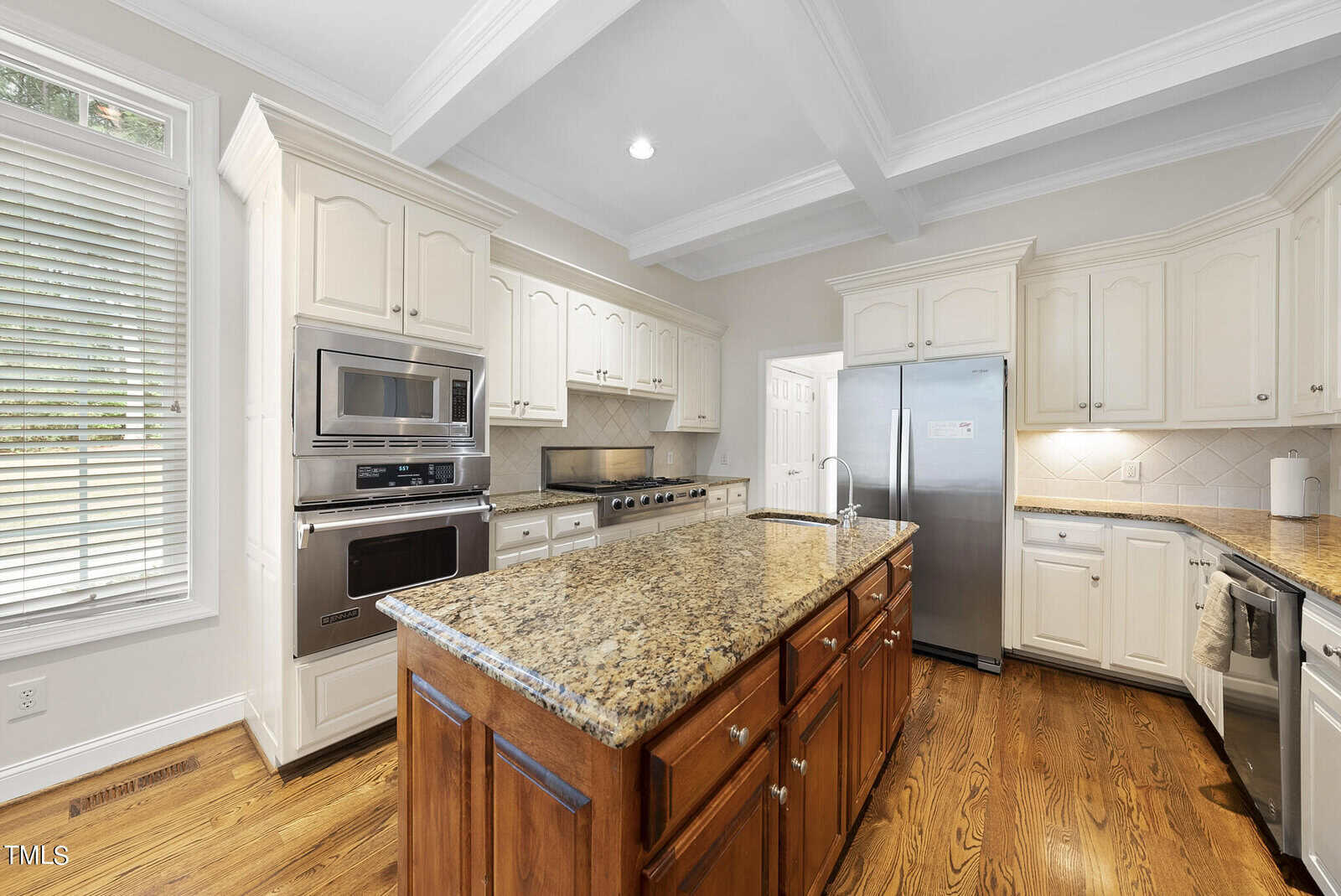 A modern kitchen with white cabinetry, stainless steel appliances, and granite countertops. An island with a wooden base and a granite top is centered. There is a large double-door refrigerator, a built-in microwave, and an oven. The floor is wood.