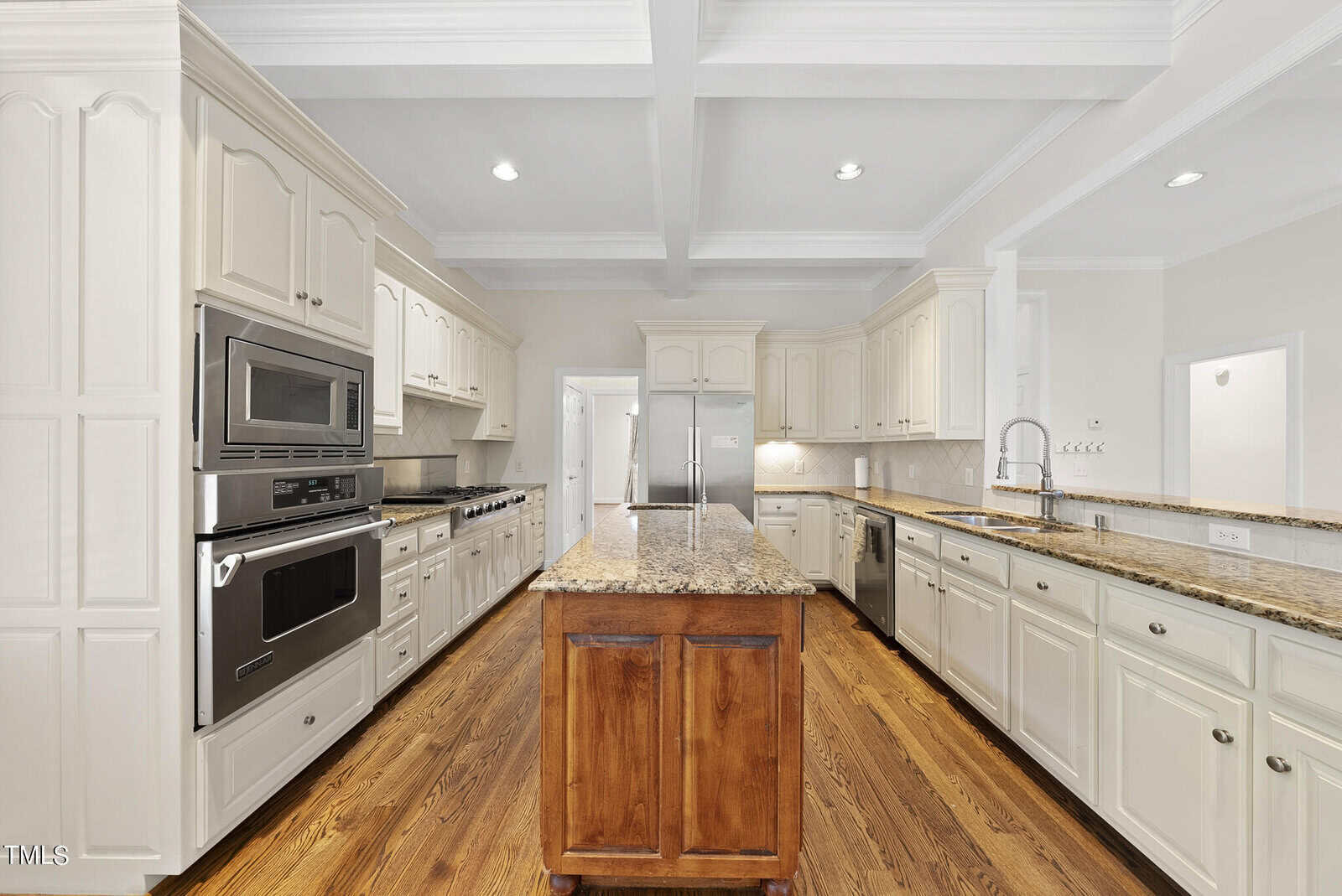 A spacious kitchen featuring white cabinetry, stainless steel appliances, and a large granite-topped island with a wooden base. The hardwood floors complement the modern design, and recessed lighting adds brightness. A doorway and a hallway are visible in the background.