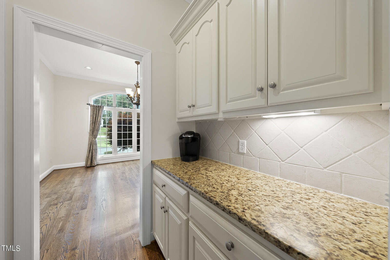 A kitchen with white cabinetry and granite countertops. A coffee maker is placed on the countertop. The kitchen opens into a bright dining area with a chandelier, wooden flooring, and large windows covered with curtains, showing a view of greenery outside.