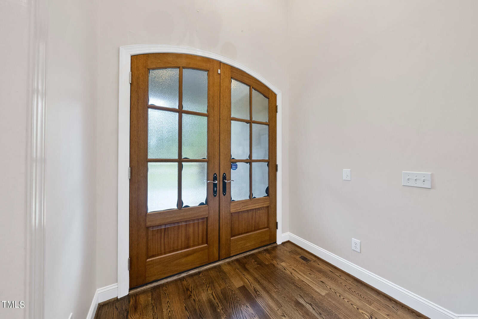 A bright interior space featuring a set of double wooden doors with glass panes, allowing natural light to enter. The doors have black handles and keyholes. The room has white walls, light switches, and a power outlet on one side, and a dark hardwood floor.