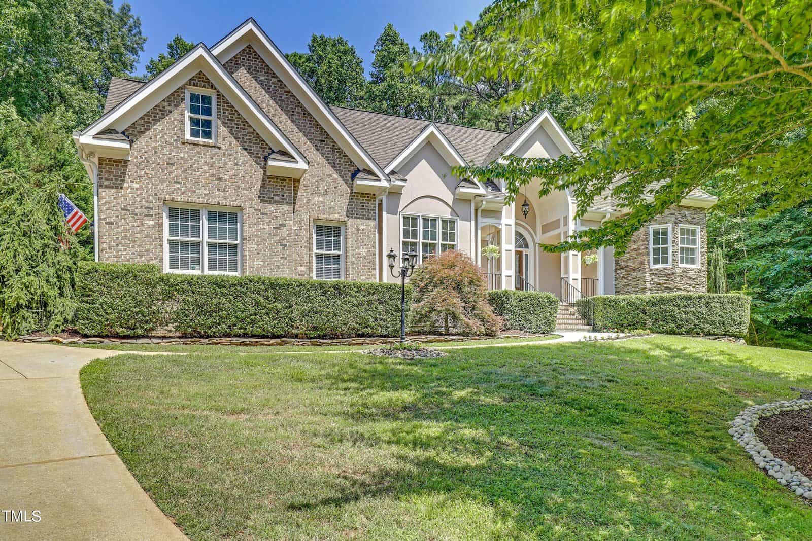 A large brick and stucco house with a gabled roof sits proudly at 2020 Silverleaf Drive, surrounded by manicured hedges and trees. A driveway leads to the front entrance, where an American flag waves near the door. The setting in Youngsville is bright under a clear blue sky.