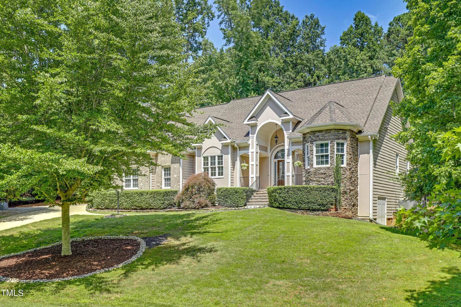A suburban house with a light-colored exterior and arched entryway surrounded by green grass and trees. The landscaping includes trimmed hedges and a mulch bed with a small bush.