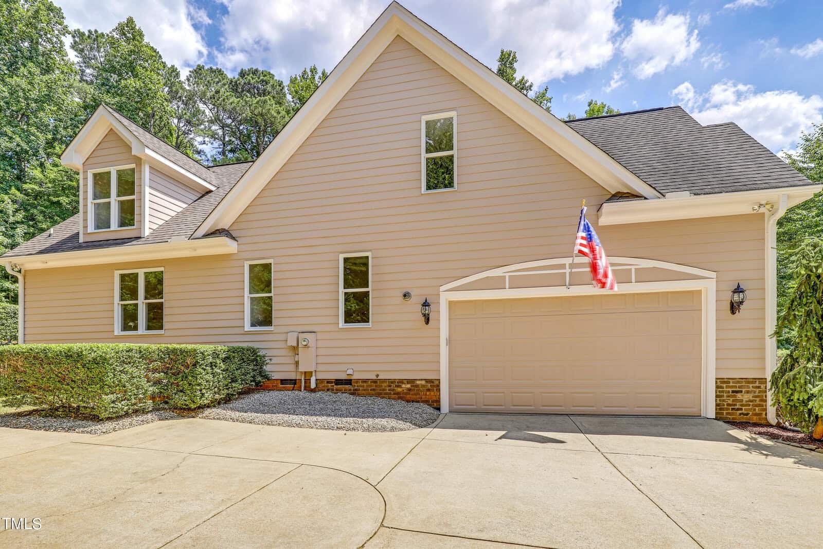 A beige two-story house with a gable roof features a two-car garage adorned with an American flag. The driveway is curved, with shrubbery on the side. Surrounded by trees, the house sits under a partly cloudy sky.