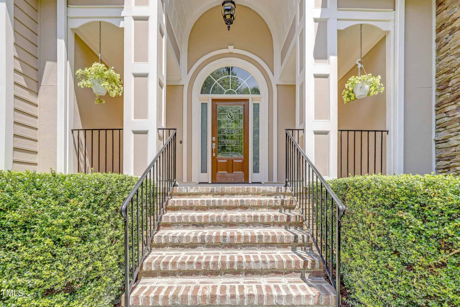 Elegant entrance of a house with brick steps leading to a wooden door with glass panels, flanked by manicured hedges and hanging flower pots under a curved stone archway.