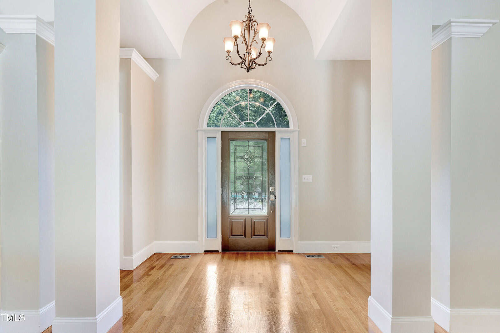 Spacious and elegant entryway featuring a wooden door with glass panels and an arched window above. The foyer has light-colored walls, a high ceiling, and a classic chandelier, with polished hardwood floors adding warmth to the space.