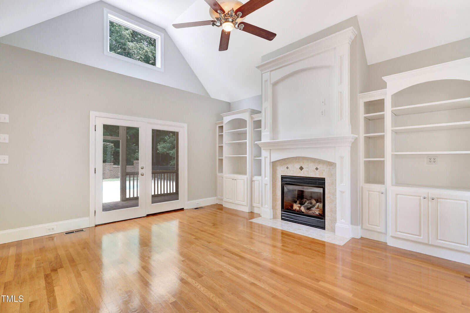 A spacious living room features a vaulted ceiling with a fan, a large white fireplace flanked by built-in shelves, and hardwood floors. A set of French doors leads to an outdoor deck, and natural light illuminates the space.