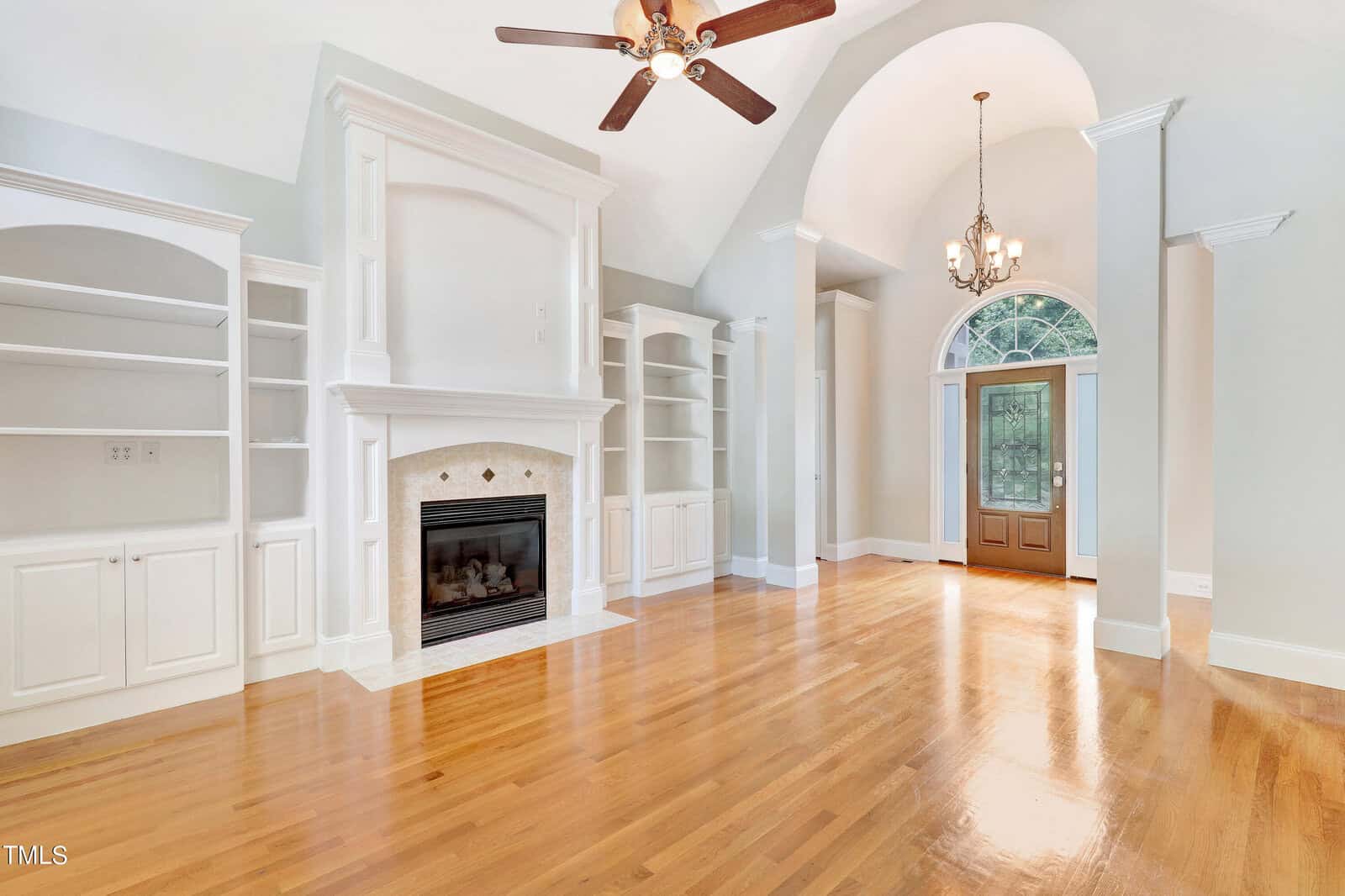A spacious living room with light wood flooring, built-in shelving, and a fireplace. The room has high, vaulted ceilings with a ceiling fan and chandelier. Large double doors with a window above allow natural light to enter.