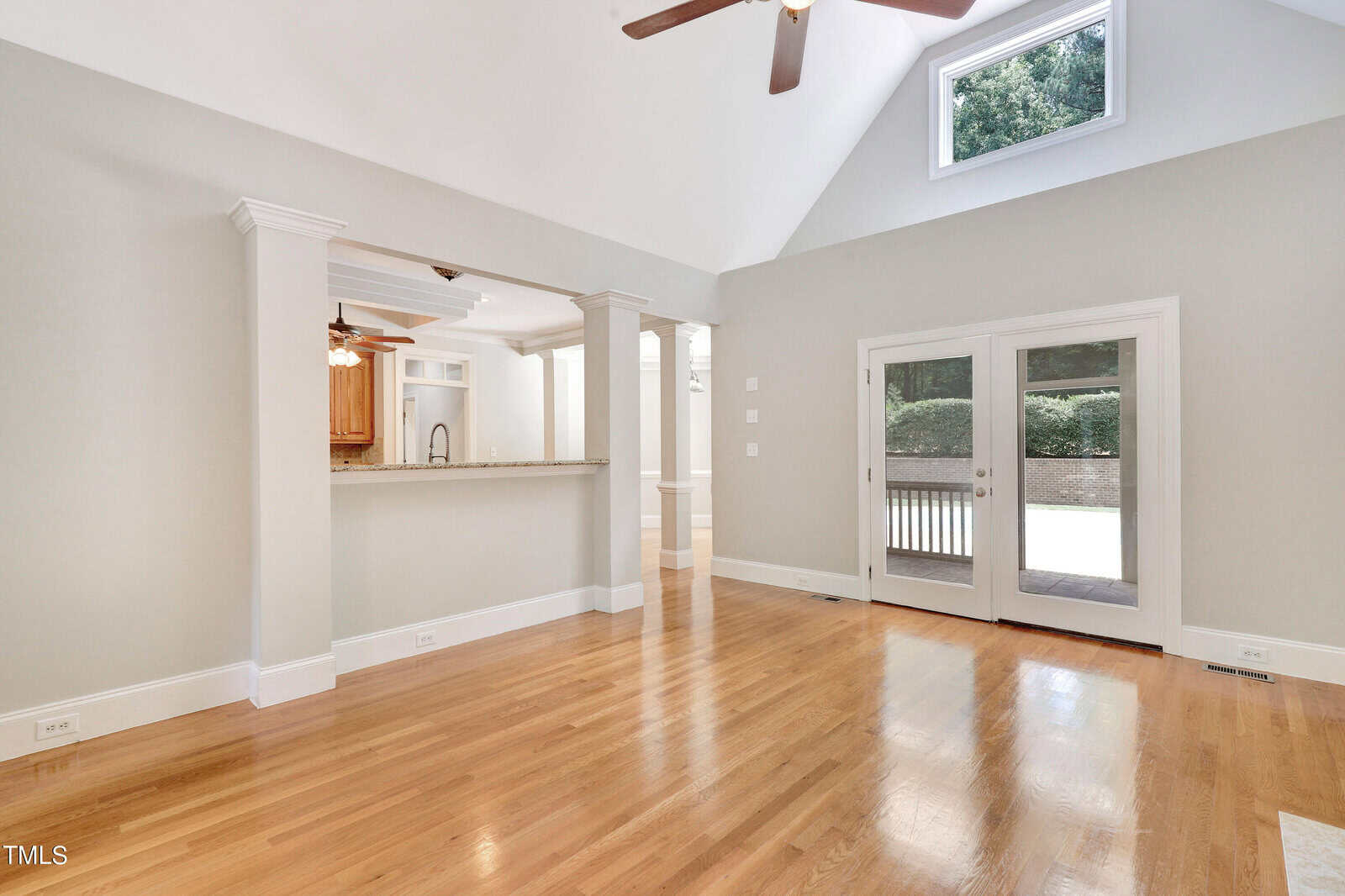 Spacious, bright room with polished hardwood floors and high, vaulted ceiling. Features a ceiling fan, large windows with a sliding glass door, and a view into a kitchen through an open partition. Walls painted light gray.