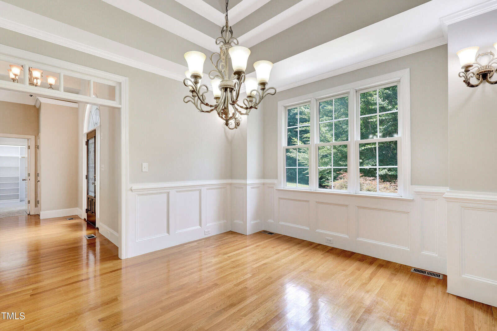 A bright, empty dining room with hardwood floors, wainscoting, and a tray ceiling. Large windows let in natural light, illuminating the elegant chandelier. A doorway leads to another room.