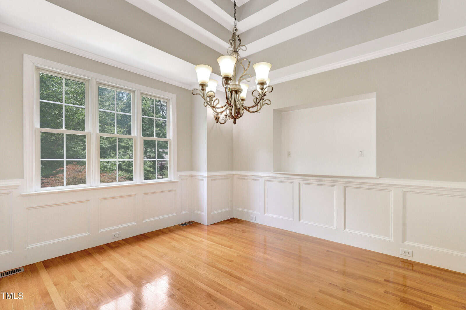 A spacious dining room with a polished hardwood floor, featuring a large window allowing natural light. The ceiling has decorative moldings, and a chandelier hangs above. Wainscoting decorates the lower half of the light-colored walls.