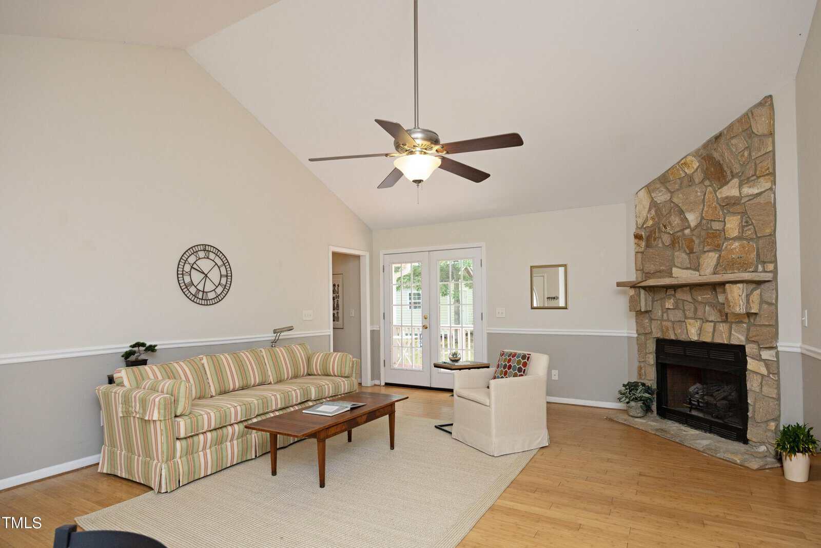 A cozy living room with a striped sofa, wooden coffee table, and a single chair arranged on a light rug. The room features a stone fireplace, a ceiling fan, and French doors leading to a patio. A decorative clock and mirror adorn the walls.