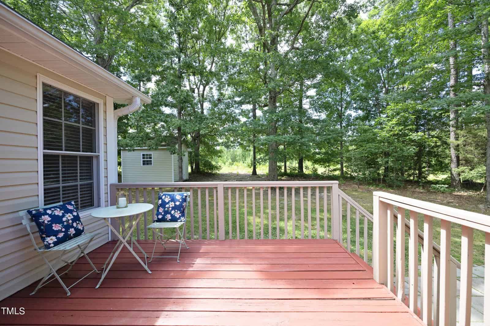A wooden deck with a round table and two chairs featuring floral cushions. The deck overlooks a backyard with lush green trees and a small shed. Sunlight filters through the canopy, creating a peaceful outdoor setting.