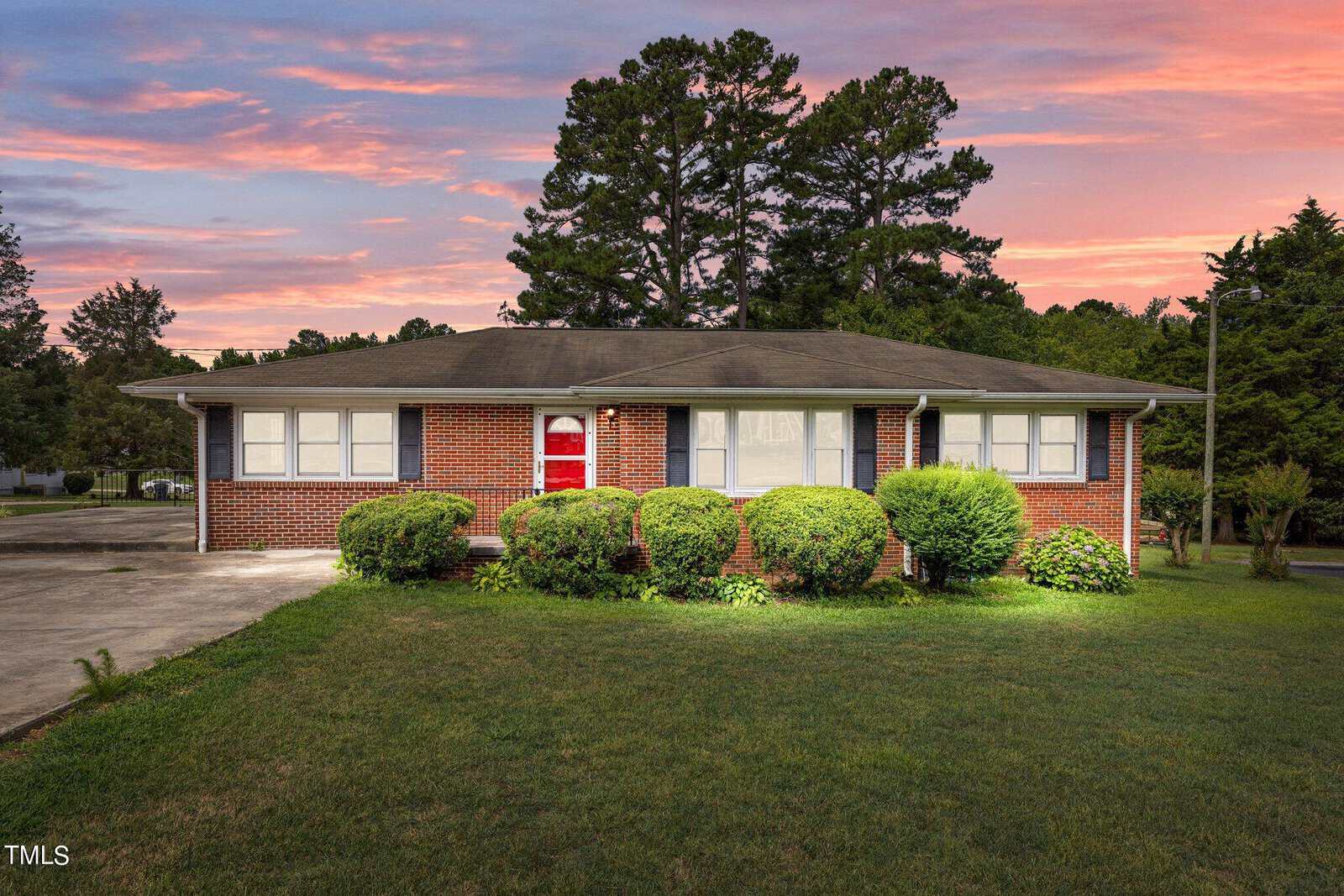 A single-story red brick house with a red door is surrounded by well-maintained bushes and a green lawn. The background features tall trees and a vibrant sunset with pink and orange hues in the sky. A driveway is visible on the left side of the house.