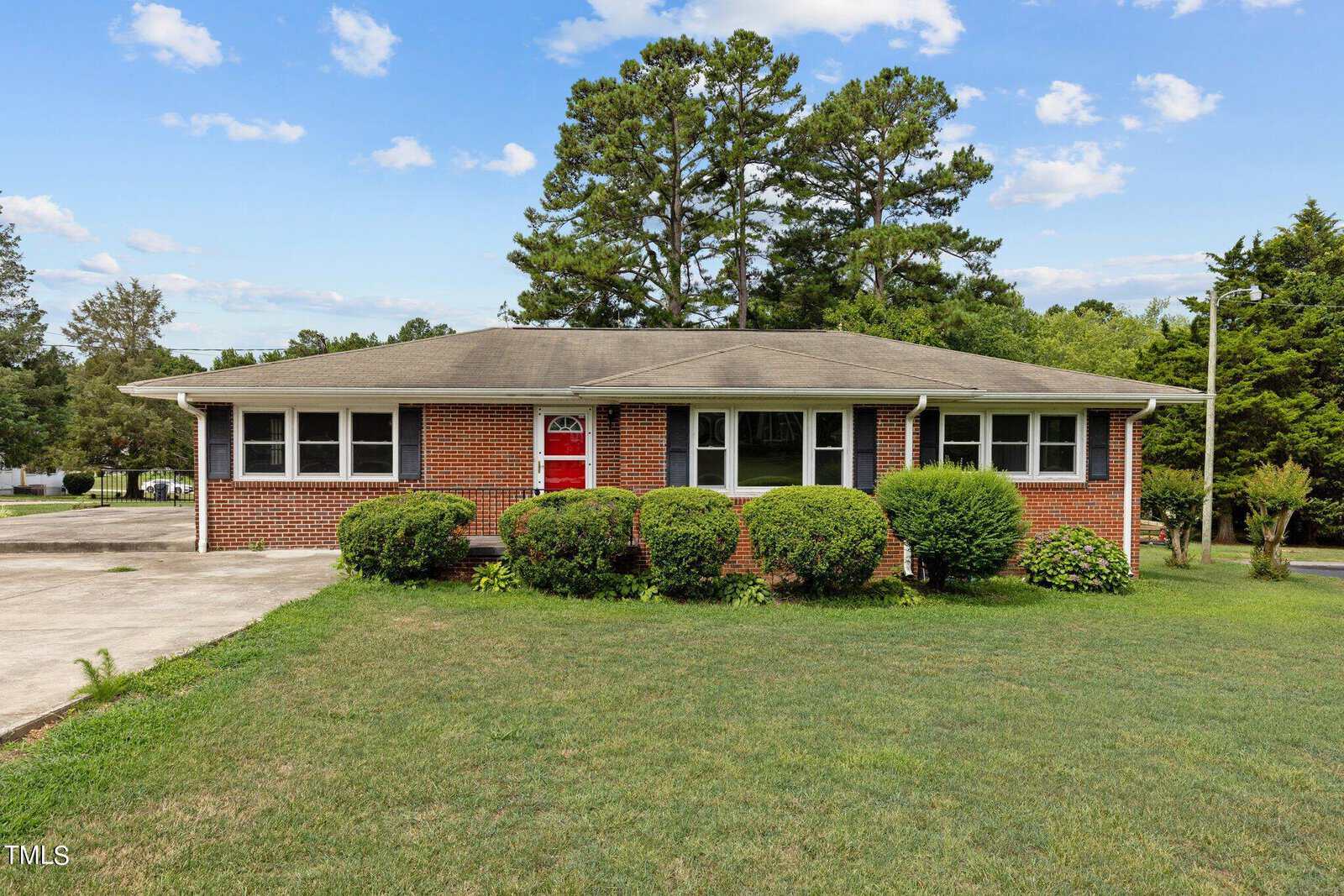 A single-story brick house with a red front door, surrounded by trimmed bushes and a well-kept lawn. There is a cement driveway to the left and trees in the background under a blue sky with scattered clouds.