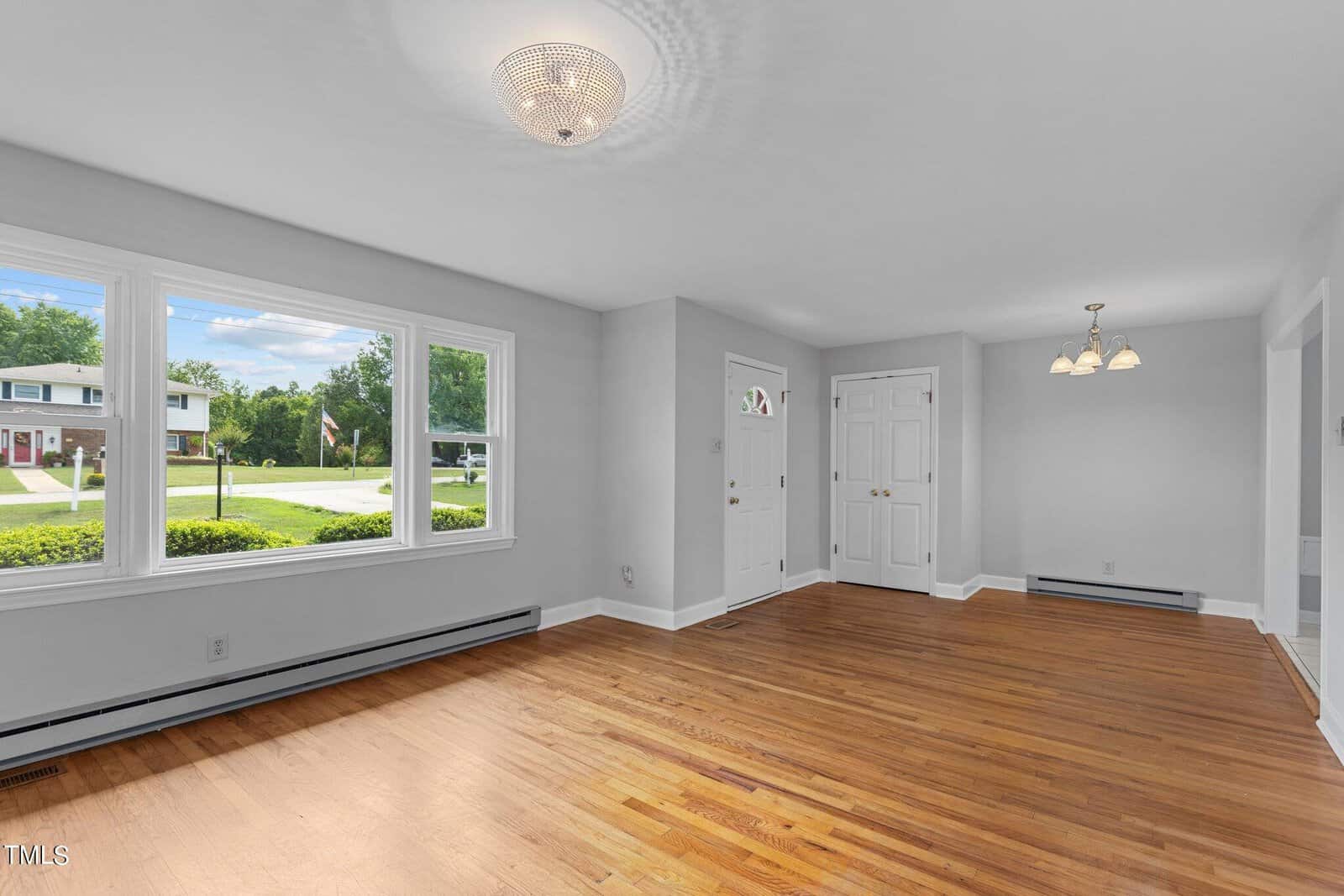 A bright, empty room with light gray walls and light wood flooring. It has large windows allowing abundant natural light. A small chandelier hangs from the ceiling. The doorway leads to a view of a residential neighborhood with green lawns and trees.