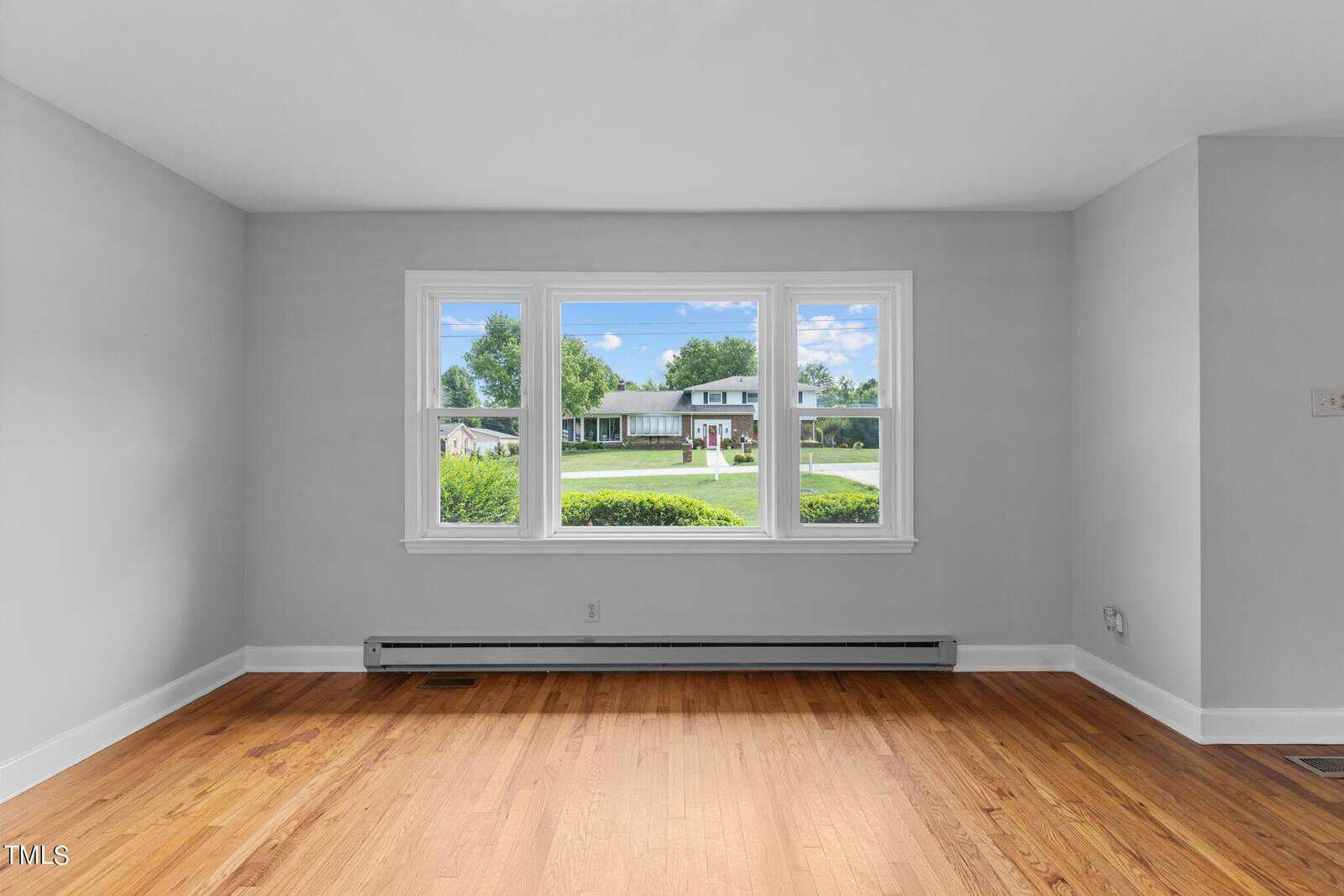 A bright, empty room with light gray walls and wooden flooring. A large window offers a view of a suburban neighborhood with green lawns and houses under a partly cloudy sky. Heating vents are visible along the base of the wall beneath the window.