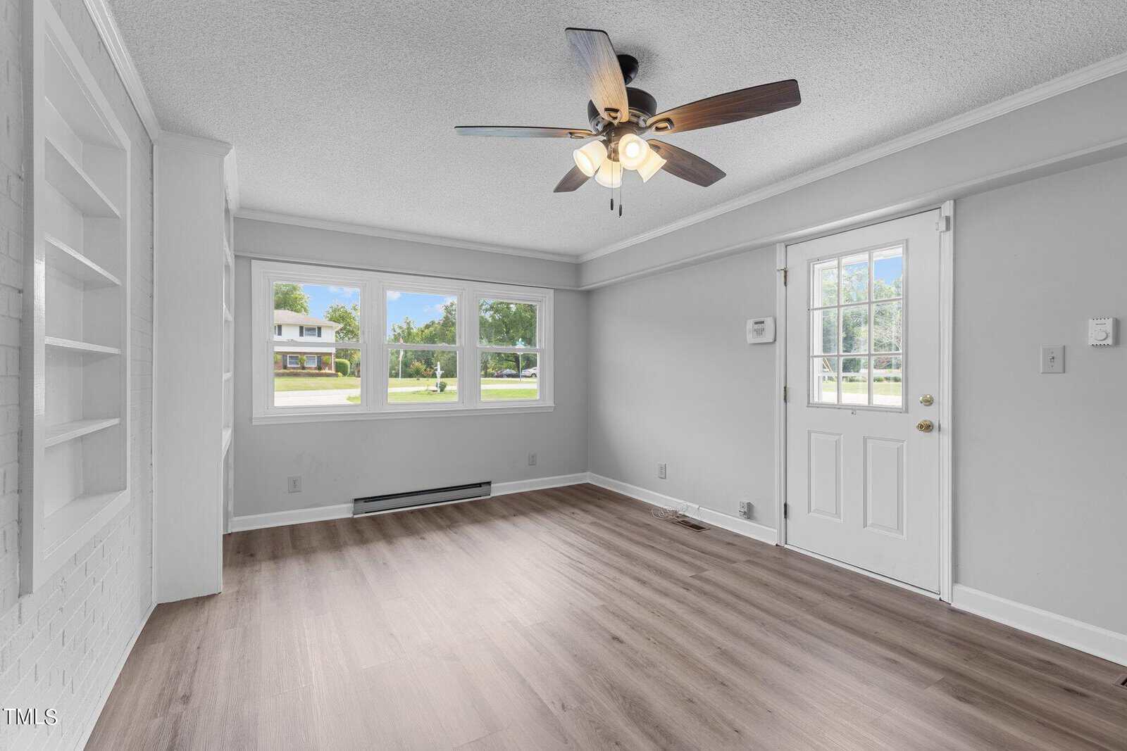 A bright, empty room with large windows, a white front door with a window, hardwood floors, and a ceiling fan with light. The room has white walls and a baseboard heater under the windows, with built-in shelving visible on the left side.