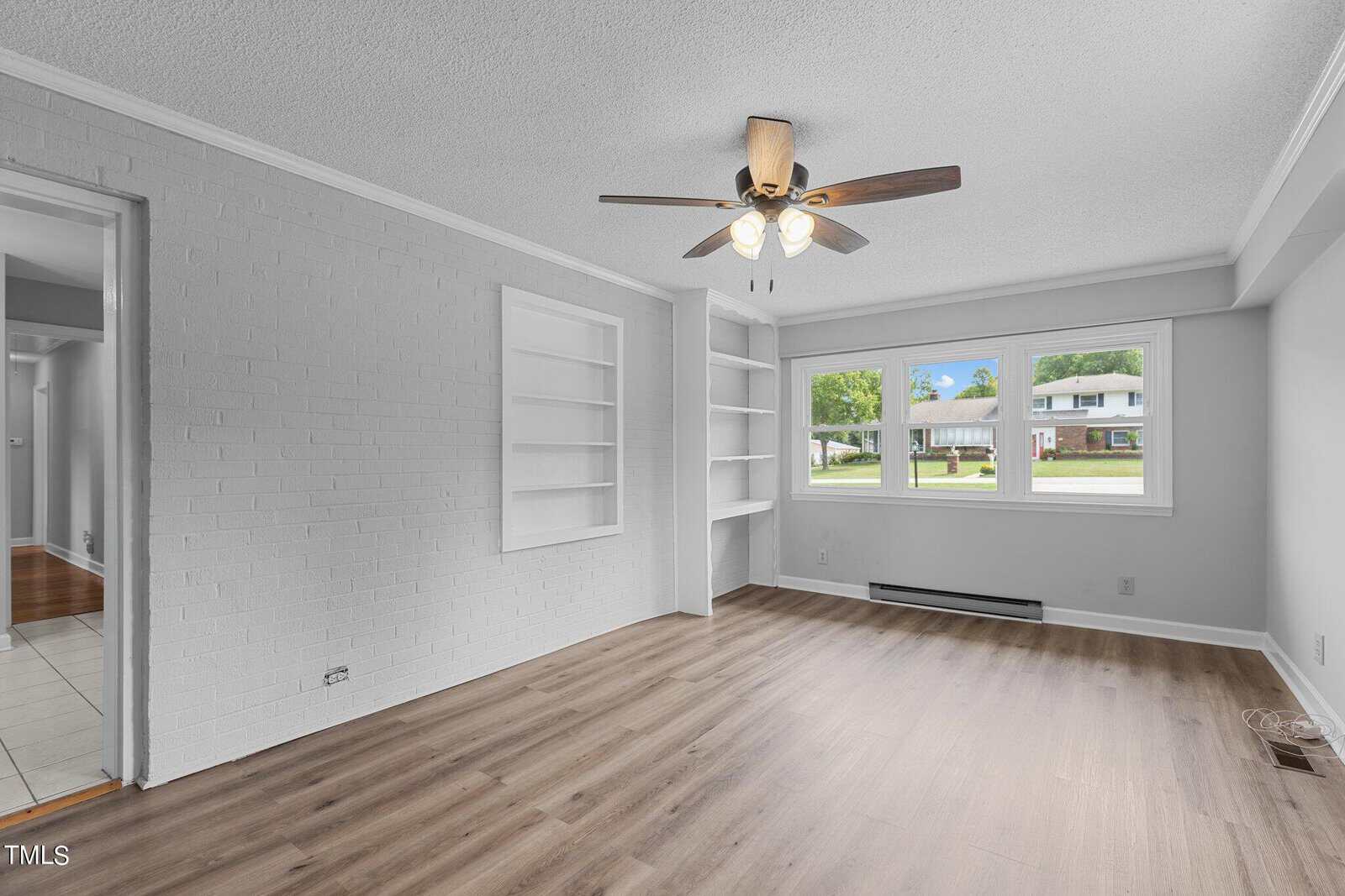 A bright, empty living room with light-colored wood flooring, white painted brick walls, built-in shelves, and a ceiling fan with lights. A large window provides a view of a green lawn and neighboring houses. An open doorway leads to another room.