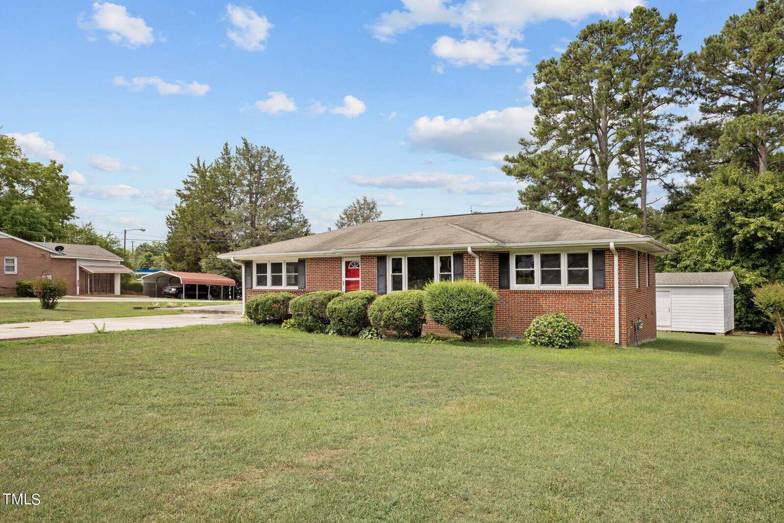 A single-story red brick house with a gray shingle roof and a well-kept lawn. Shrubs line the front of the house. There are trees in the background, and a driveway extends to the left, leading to a carport. The sky is blue with scattered clouds.