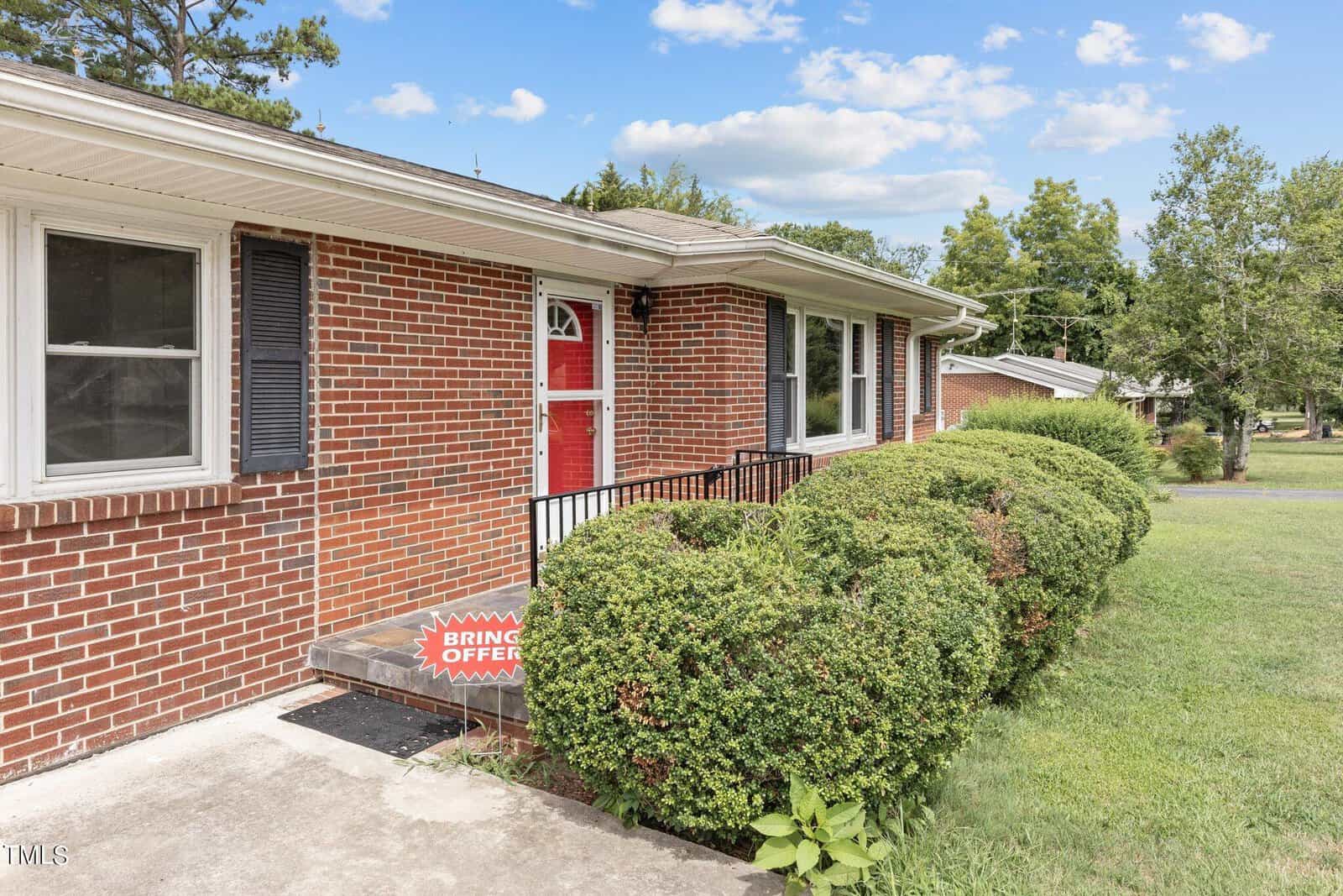 A single-story brick house features a red front door with adjacent windows. Well-maintained hedges line the front yard, and a sign near the steps reads "Bring Offers." The background includes lush green trees and a partly cloudy sky.