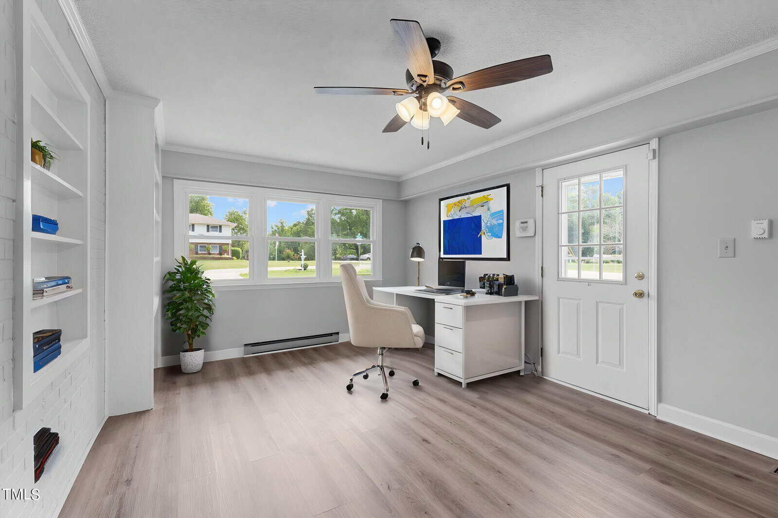 A bright home office with a large window displaying a green yard outside. There's a white desk with a computer, a beige chair, and a potted plant in the corner. Open shelving on the left contains books and decor items. A ceiling fan with lights hangs above.