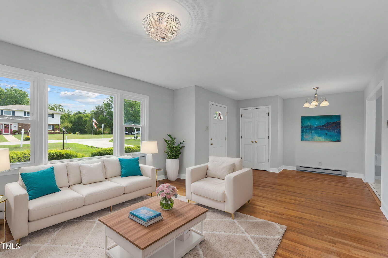 A bright living room with light gray walls and wood flooring features a white couch and matching armchair, both with teal throw pillows. A large window lets in natural light, and a coffee table with a floral centerpiece is centered on a beige rug. A chandelier hangs from the ceiling.