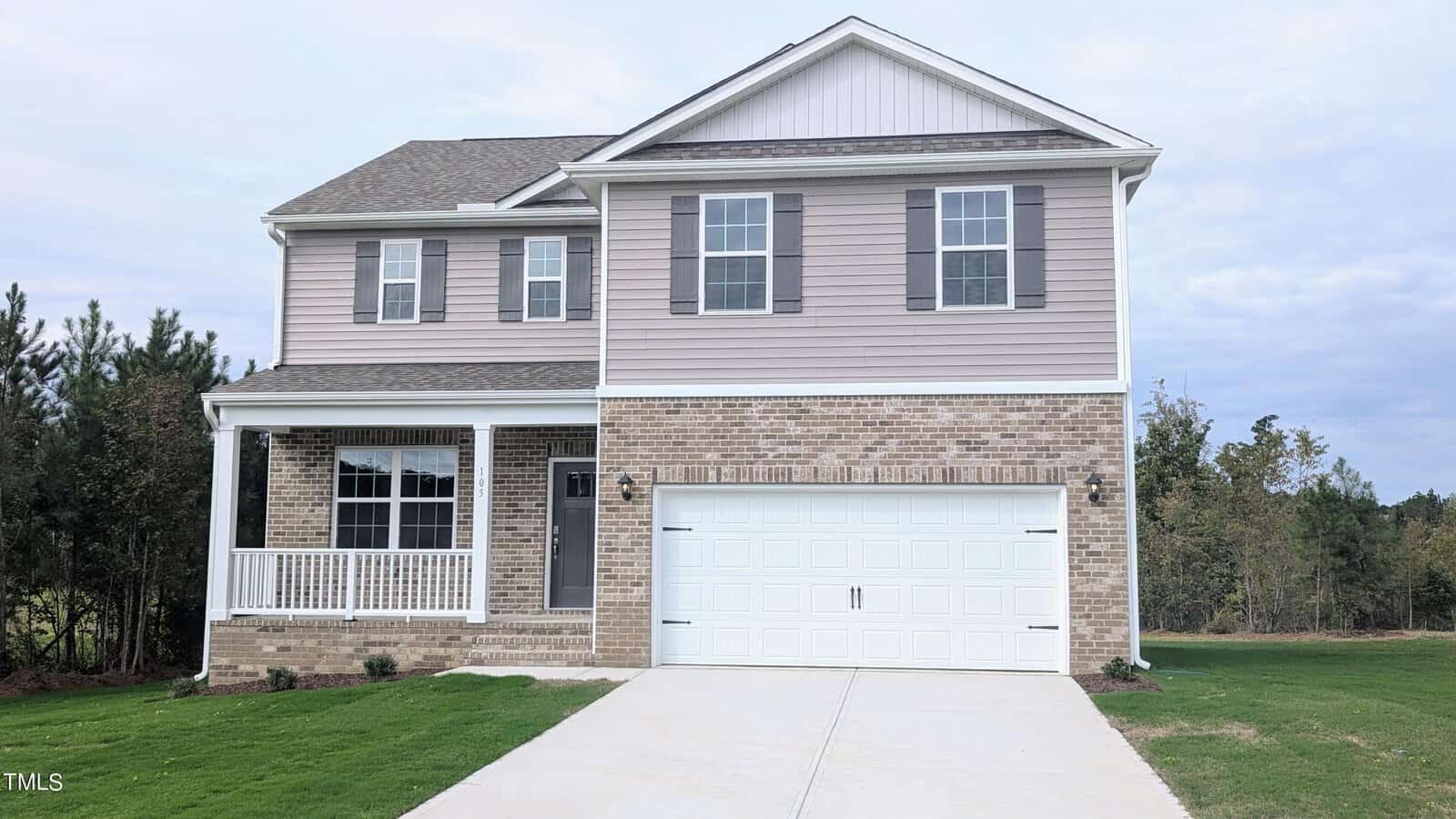 A two-story suburban house with beige siding and brick facade, gray shutters, a white front porch, and a double garage, set on a green lawn with trees in the background under a cloudy sky.