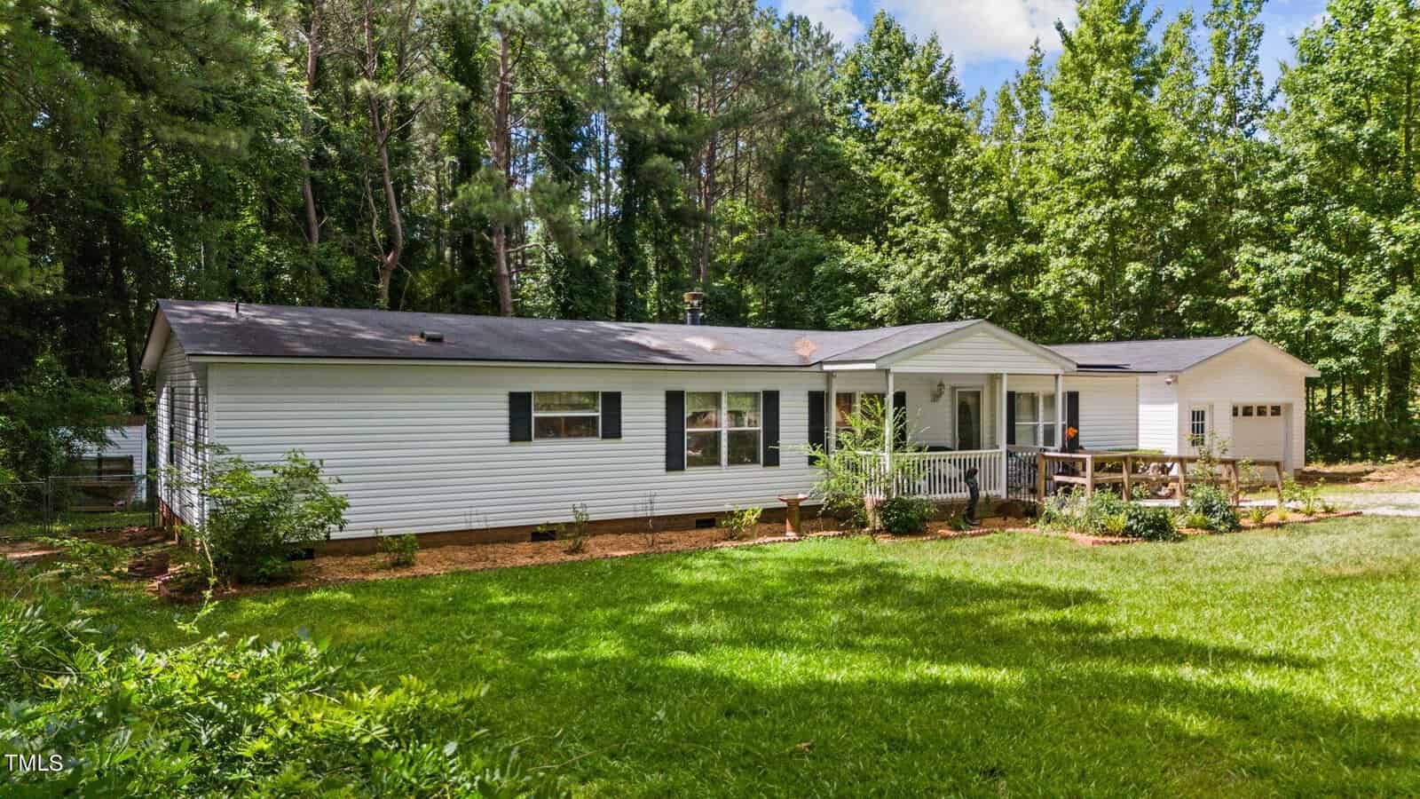 A white, single-story house with a dark roof, located in a lush, green yard surrounded by tall trees. The house has a small porch with a railing and an attached garage. The sky is partly cloudy.