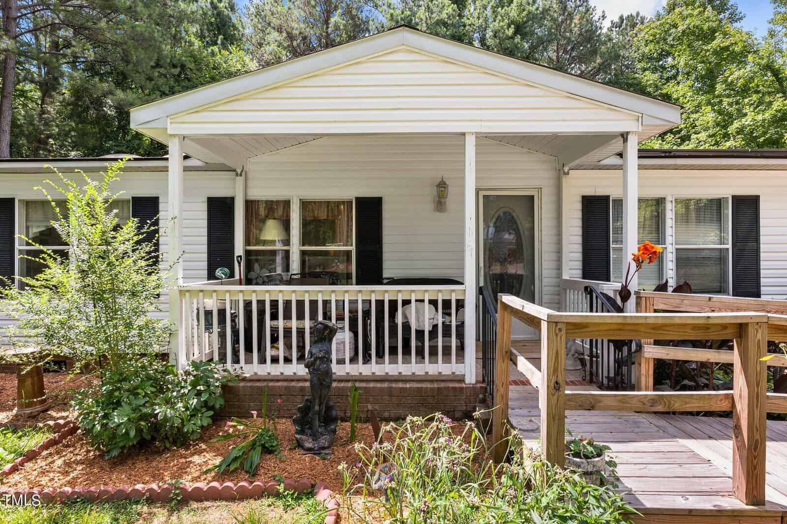 A white mobile home features a covered porch with white railings and a ramp leading up to the entrance. There's a statue of a dog in the garden on the left, surrounded by greenery and mulch. Trees can be seen in the background.