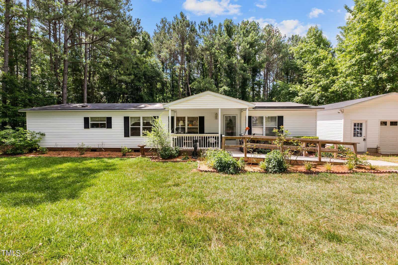 A white single-story house at 3536 Bruce Garner Road in Wake Forest features a ramp leading to a porch, nestled in a yard with lush green grass. It's surrounded by tall trees under a partly cloudy sky, and there's a garage on the right side.