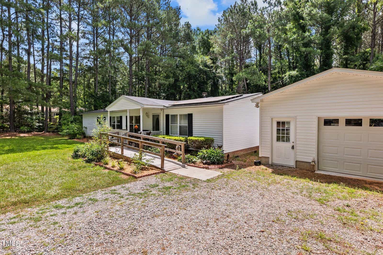 Single-story white house with a wheelchair ramp in front, surrounded by trees and greenery. A single-car garage is attached, and a gravel driveway leads to the entrance.