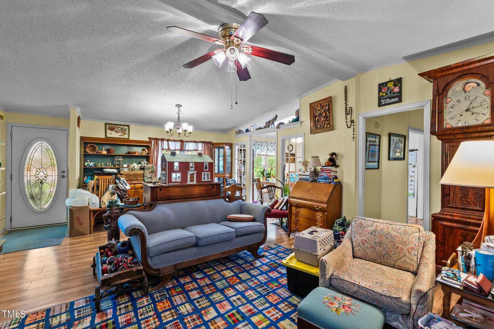 A cozy living room with a blue couch, patterned rug, and various antique furniture. On the left, there's a wooden cabinet and a globe. A ceiling fan hangs above, and natural light fills the room through windows and an open door.