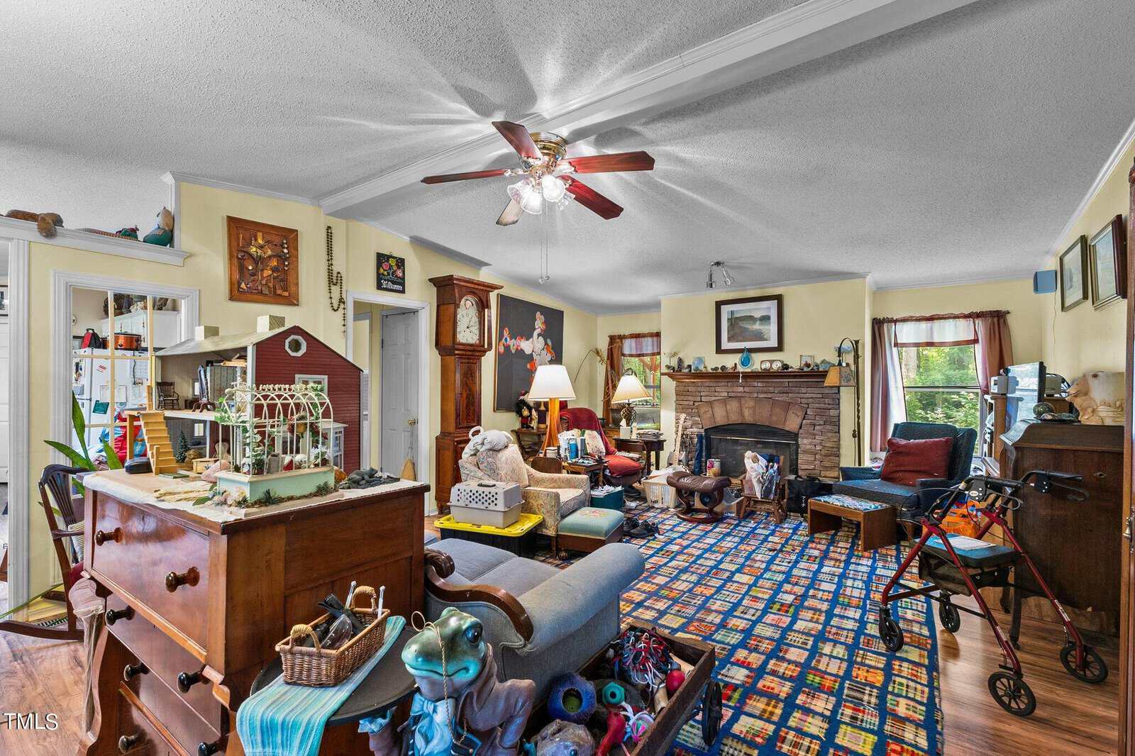 A cluttered living room filled with various items, including a wooden chest, model house, toys, and a walker. The room has a patterned carpet, white ceiling fan, fireplace, and a mix of eclectic decor on the walls. Chairs and tables are scattered around.
