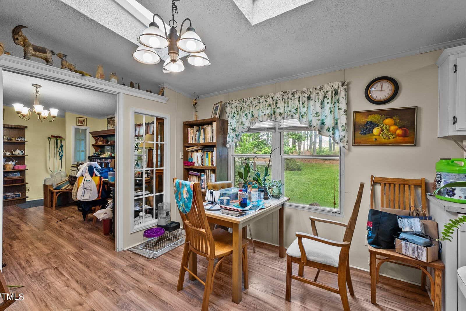 A cozy dining area with wooden floor and furniture. The room is decorated with floral curtains and various decorations on the shelves. A table and chairs are set by a window showcasing a view of a green yard outside. Natural sunlight fills the space.