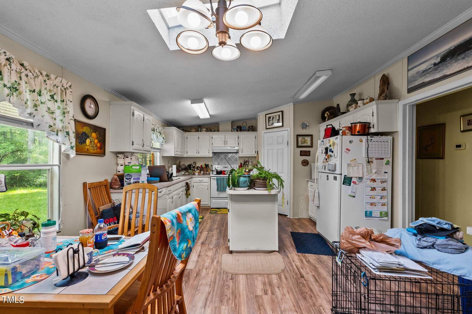 Kitchen with wooden floor, skylight, and white cabinets. Central island with potted plants. Dining table with chairs on the left. Fridge covered with magnets and papers. Various items and artwork are scattered throughout the space. Bright natural light.