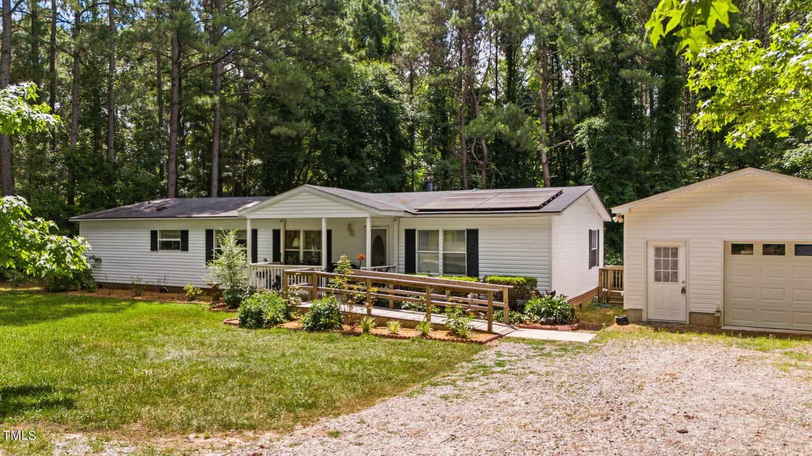 A single-story, white house with a ramp entrance surrounded by lush greenery and tall trees. Next to it is a detached garage. A gravel driveway leads up to the home, and the front yard has well-maintained shrubs and grass.