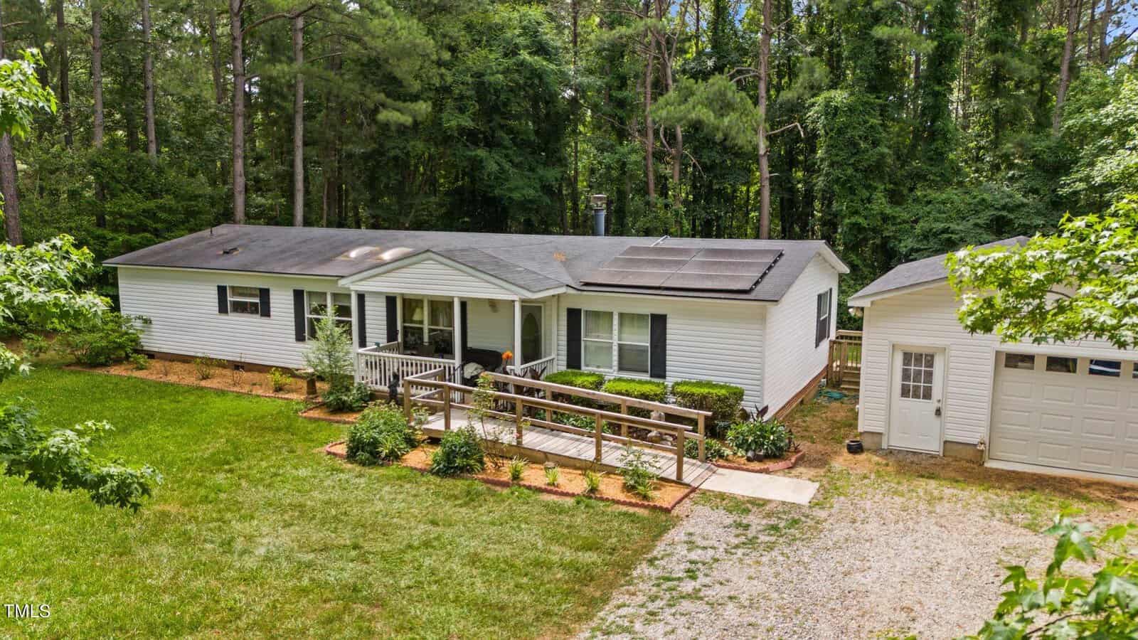 A white single-story house with a ramp leading to the front porch, surrounded by trees and greenery. A detached white garage is on the right, with a gravel driveway extending from it. Solar panels are visible on the house roof.