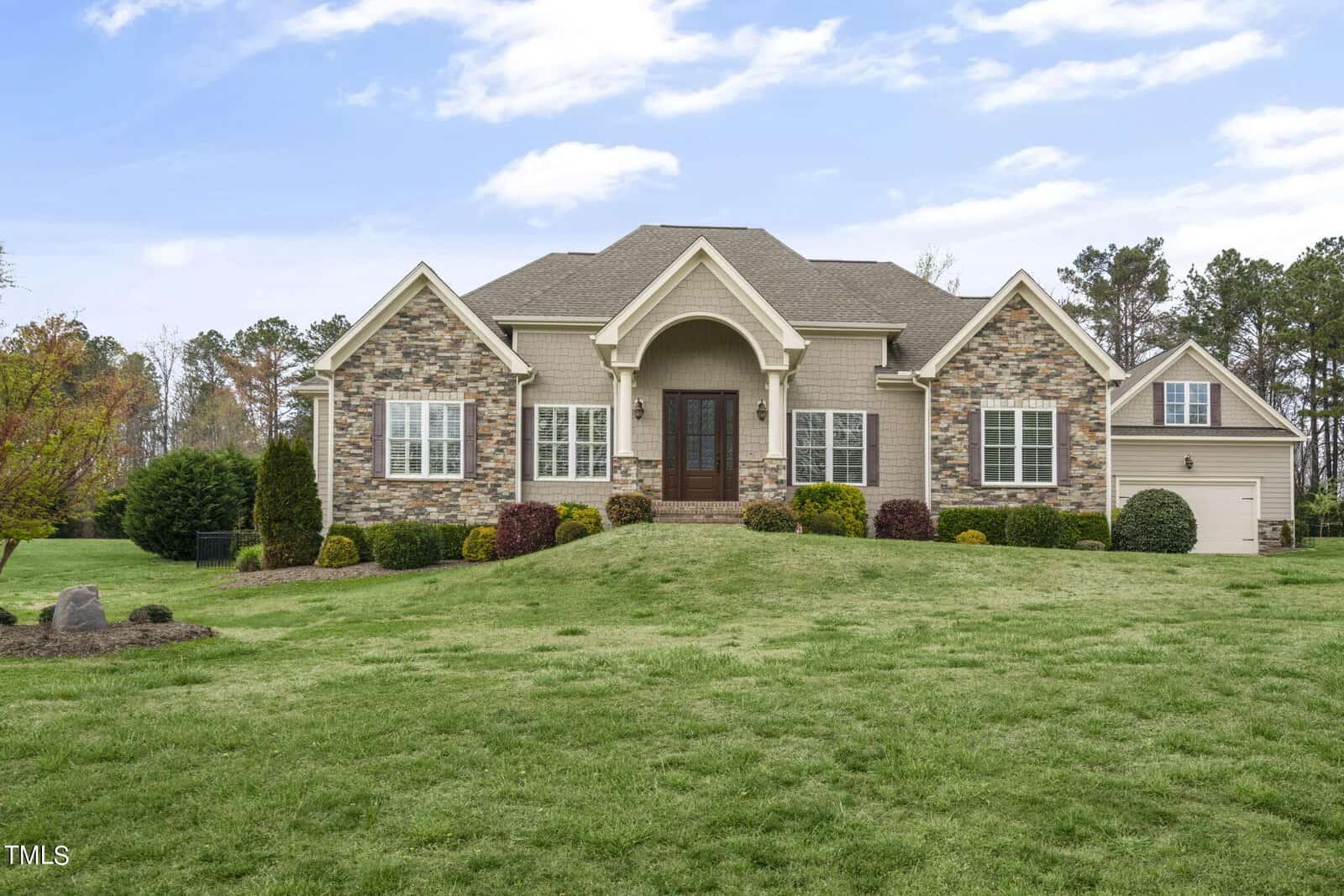 The spacious house at 1103 Dovefield Lane in Youngsville boasts a stone and beige exterior set against a lush green lawn. Its gable roof, arched entryway, and double door add charm. Encircled by shrubs and trees, the property features a garage to the right beneath a partly cloudy sky.