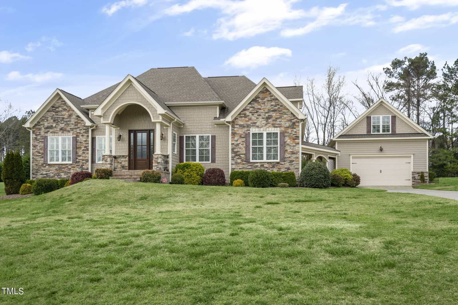 A large, modern house with a stone facade, gabled roof, and a manicured lawn. It features a front porch and a detached two-car garage. The sky is partly cloudy, and trees are visible in the background.
