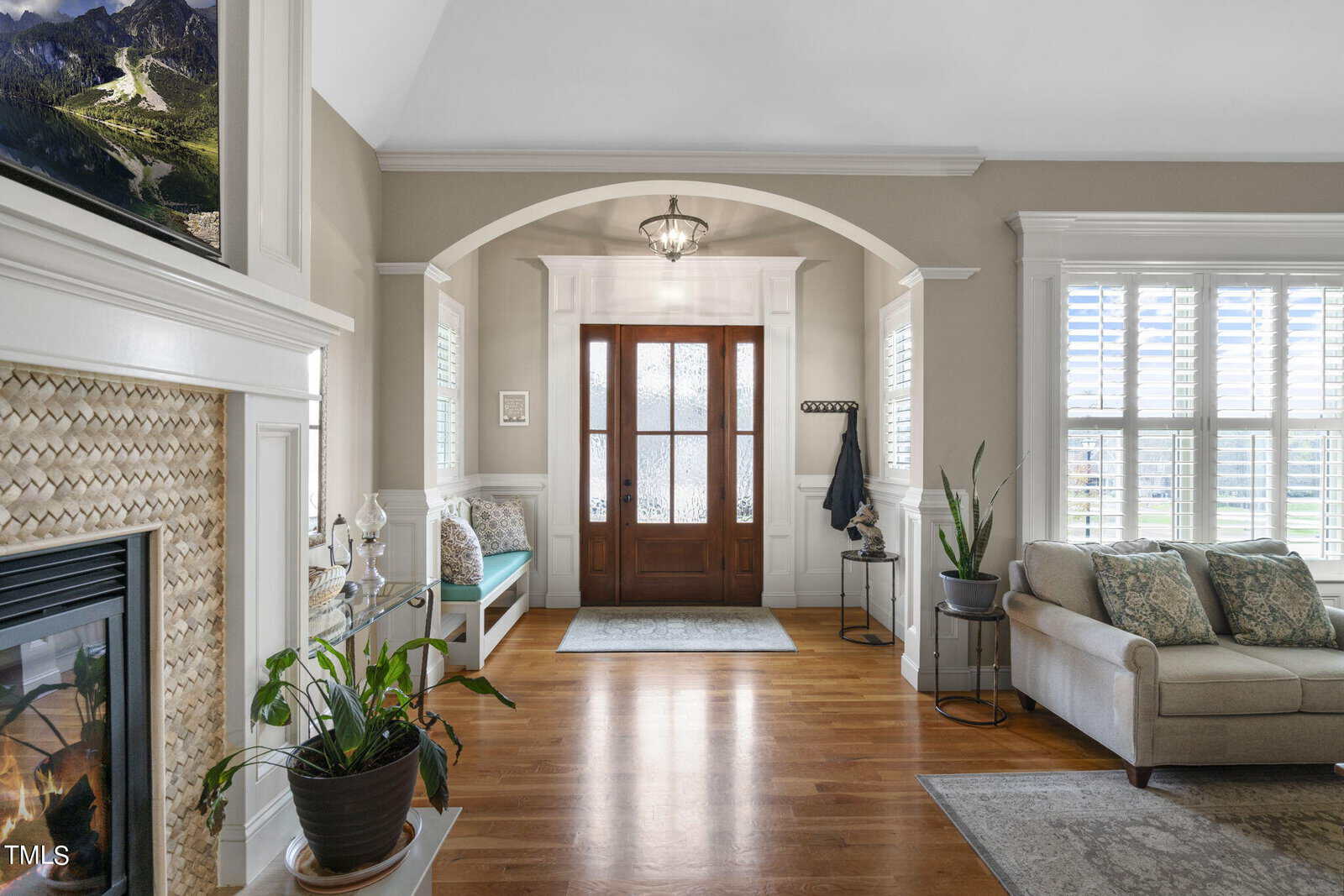 Spacious living area with a wooden floor, featuring a cozy sofa, a fireplace, and several potted plants. Sunlight streams through large windows with shutters. A wooden front door with glass panels is framed by an archway under a hanging light fixture.