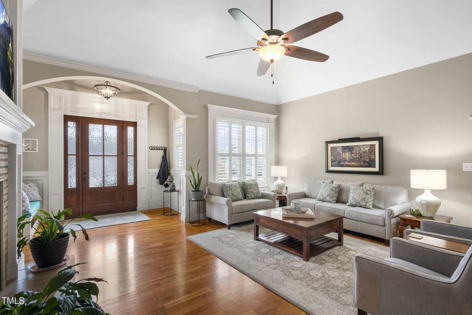 Spacious living room with hardwood floors, light gray sofas, and armchairs. A ceiling fan hangs above. There's a wooden coffee table, a large painting on the wall, and plants by the entrance with a coat rack and decorative light fixture.