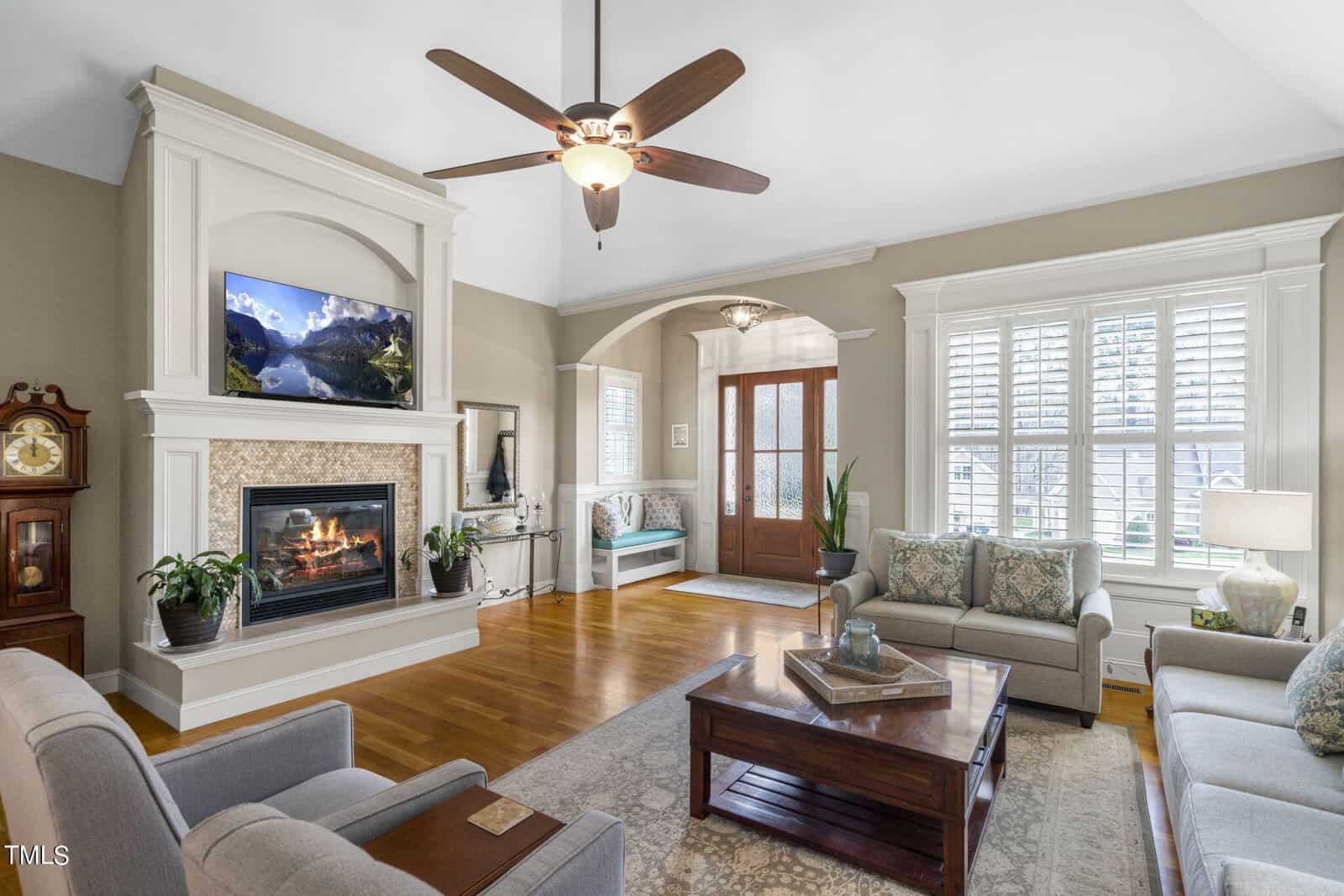 Spacious living room with hardwood floors, a ceiling fan, and a cozy fireplace. A TV is mounted above the fireplace. The room features comfortable seating, a coffee table, and large windows with shutters, offering a bright and inviting atmosphere.