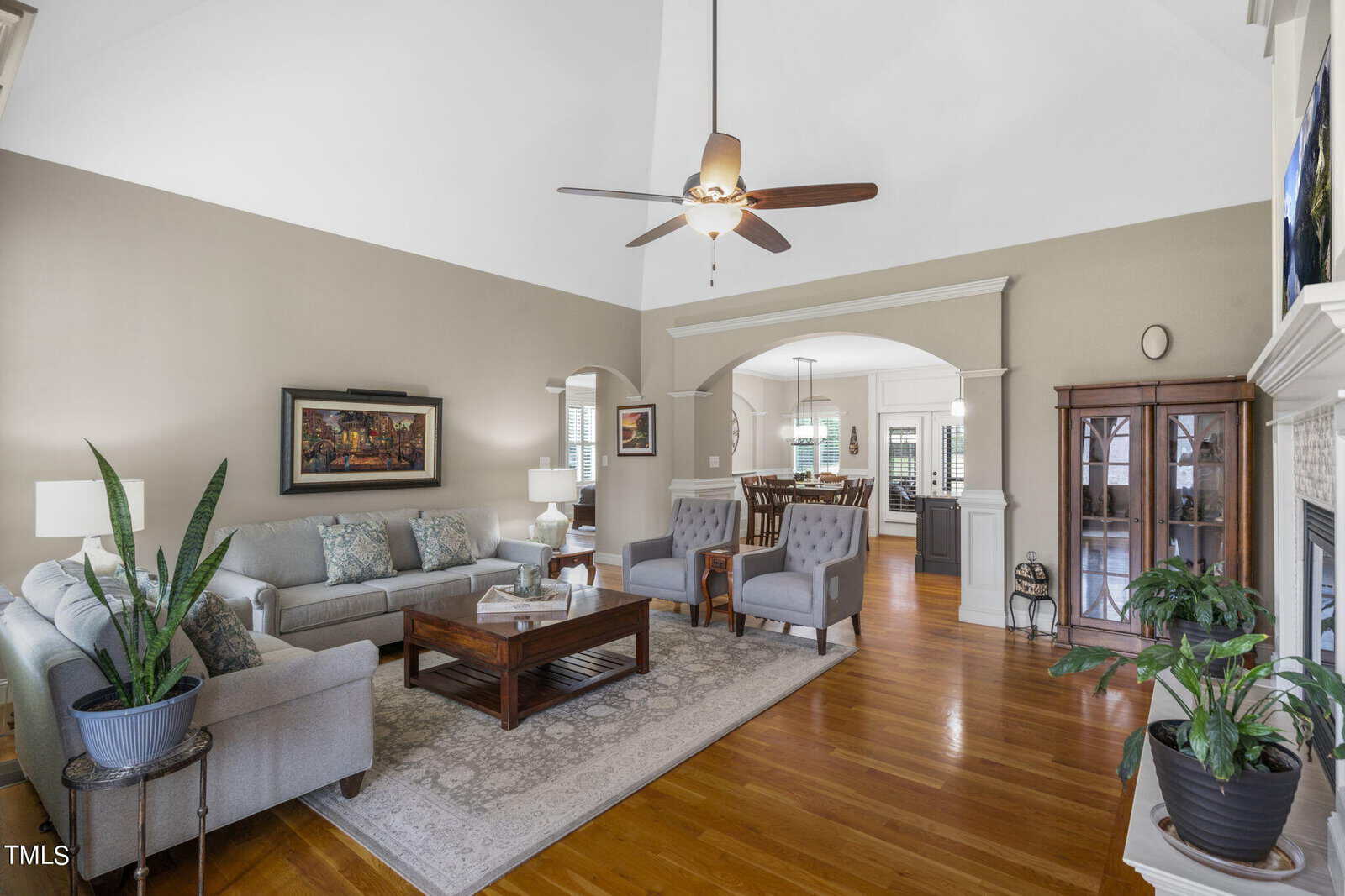 Bright living room with vaulted ceiling, chandelier fan, hardwood floors, and gray walls. Includes a gray sofa, two armchairs, coffee table, and potted plants. An archway leads to the dining area with a table and chairs in the background.