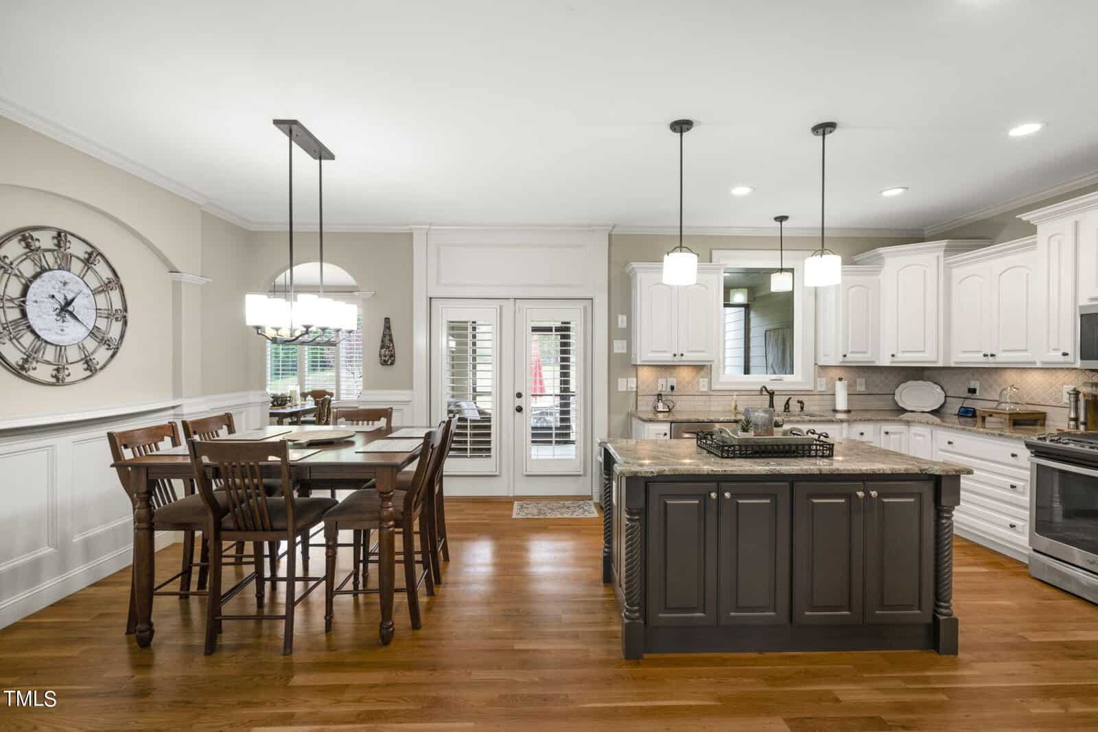 A modern kitchen and dining area with wooden floors. The kitchen has white cabinets, a central island with dark cabinets, and pendant lights. A round clock adorns one wall, and a dining table with chairs is nearby, next to a door with glass panes.