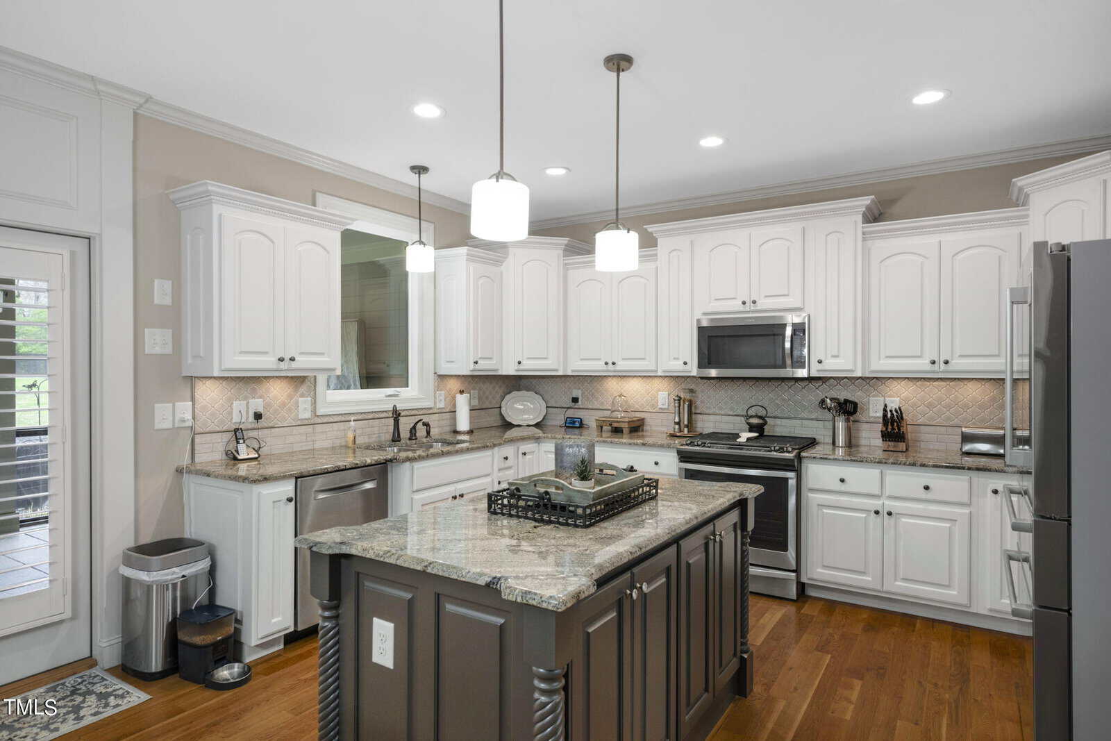 A modern kitchen featuring white cabinets, a large island with a granite countertop, stainless steel appliances, and wooden flooring. Pendant lights hang over the island, and a window above the sink provides natural light.