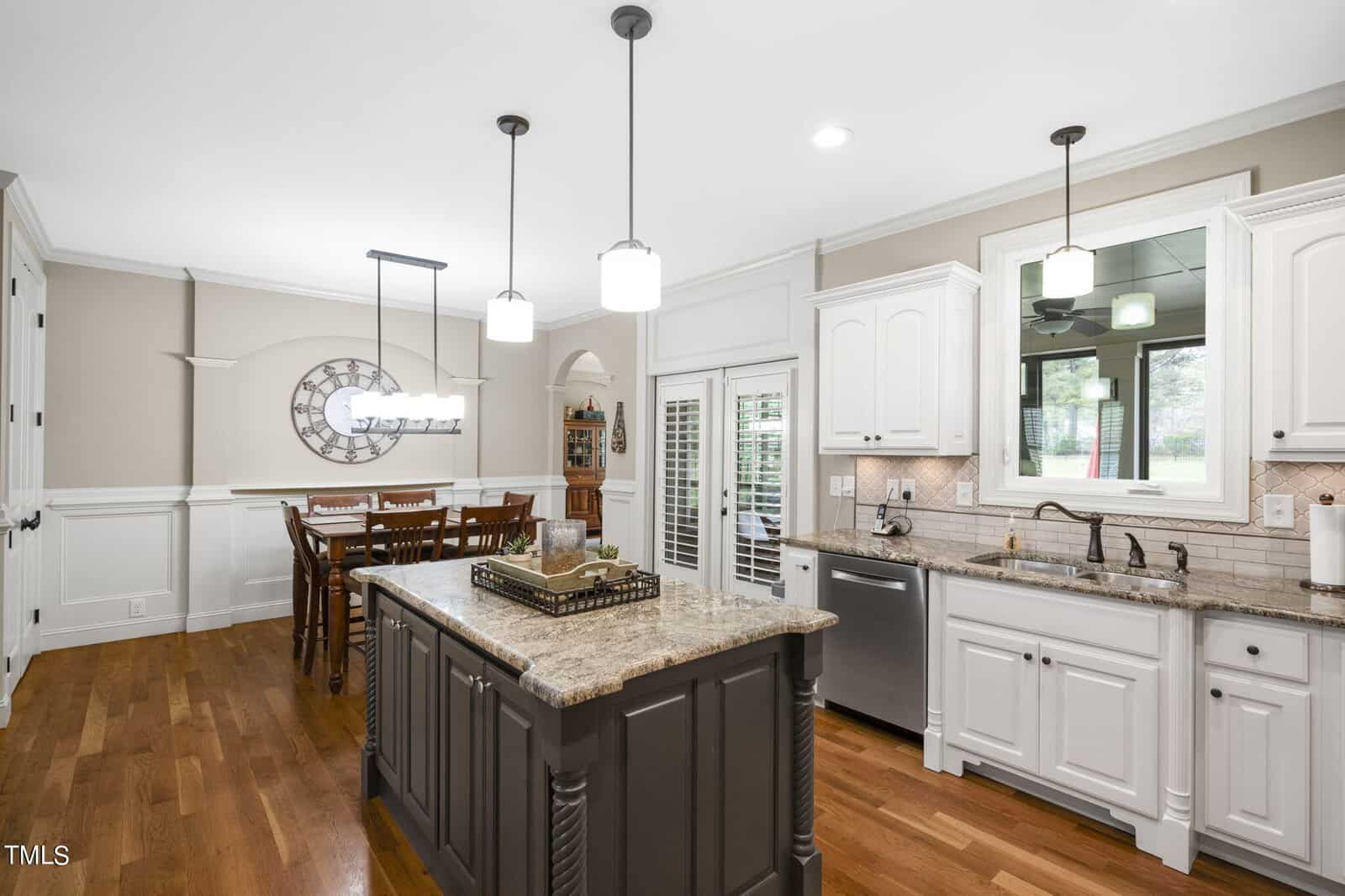 A spacious kitchen with an island and granite countertops. White cabinets line the walls, and the room features hardwood floors. Open dining area with a wooden table and six chairs, large window on the right providing natural light.