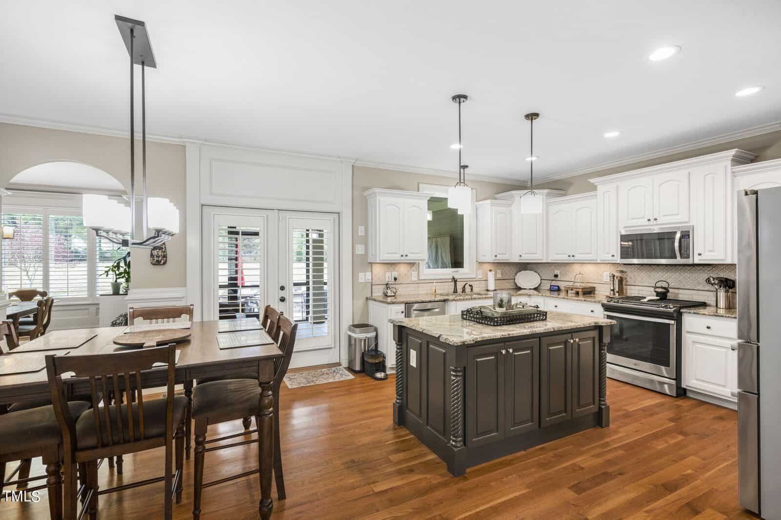 A modern kitchen with white cabinets and stainless steel appliances. An island with a granite countertop is in the center, surrounded by wooden chairs. The room is well-lit with pendant lights and has a dining area with a large wooden table.