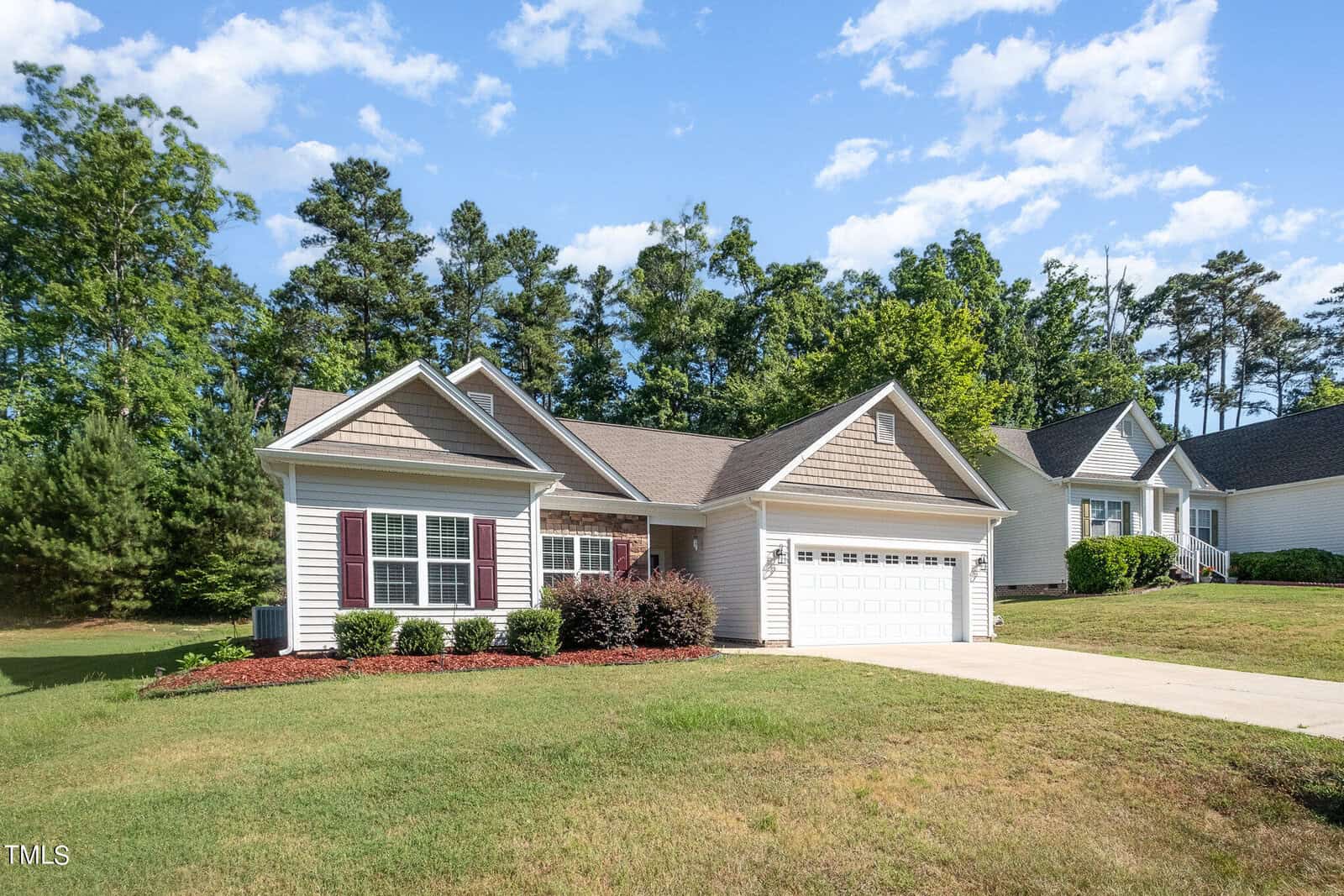 A single-story house with a light-colored exterior and attached garage in a suburban setting. The lawn is well-maintained with shrubs lining the front. Tall trees are in the background under a partly cloudy sky.