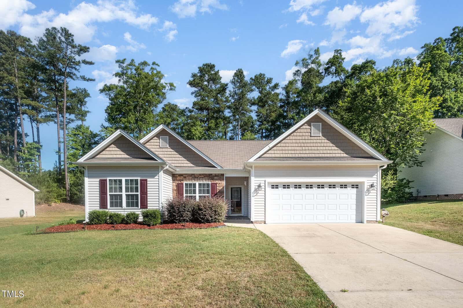 A single-story house with a light-colored facade and a two-car garage is surrounded by a well-maintained lawn. The background features tall trees and a blue sky with scattered clouds.