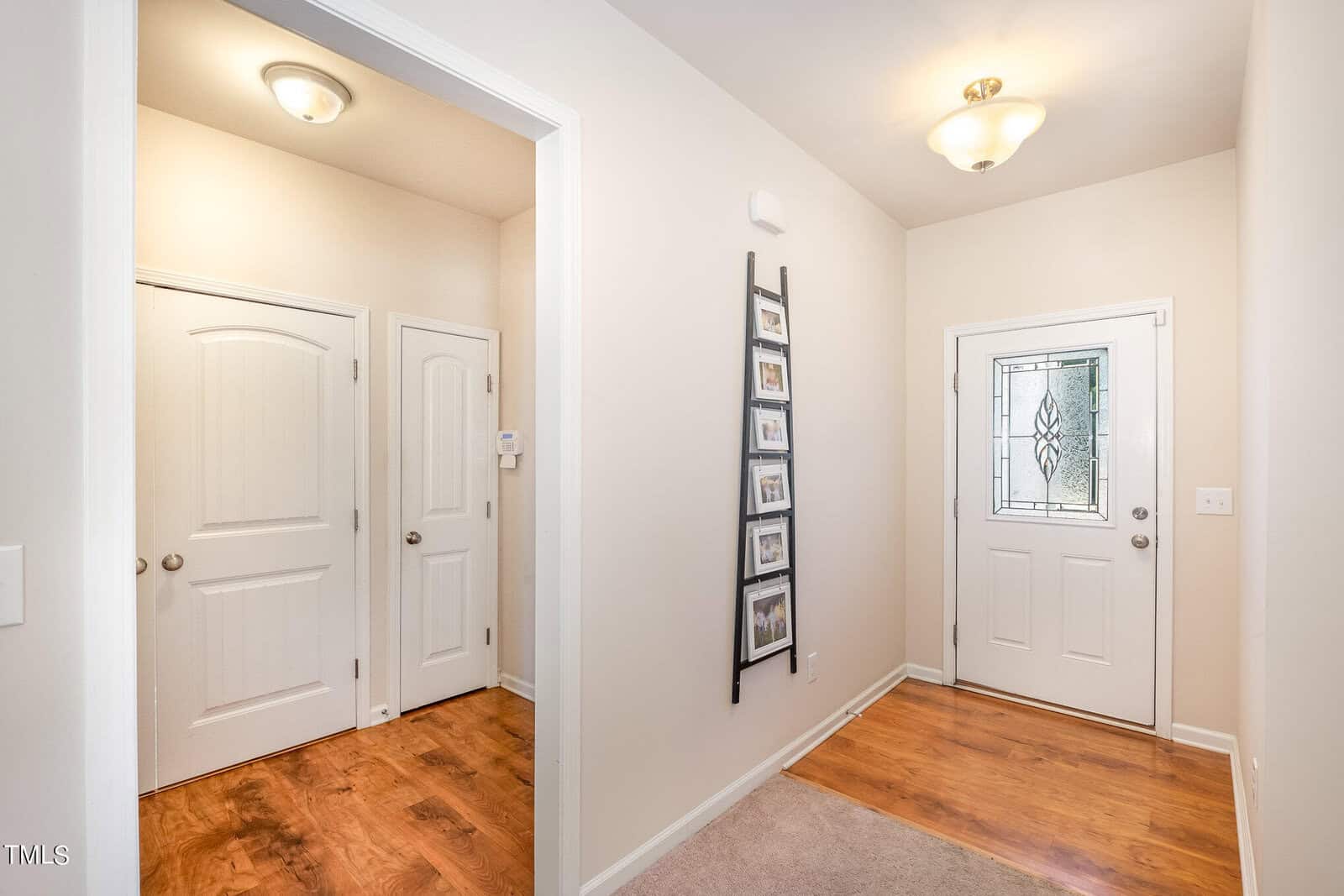 A bright hallway with a wooden floor, white walls, and two closed white doors on the left. A decorative ladder with framed photos leans against the wall. A door with patterned glass is at the end, under a ceiling light fixture.